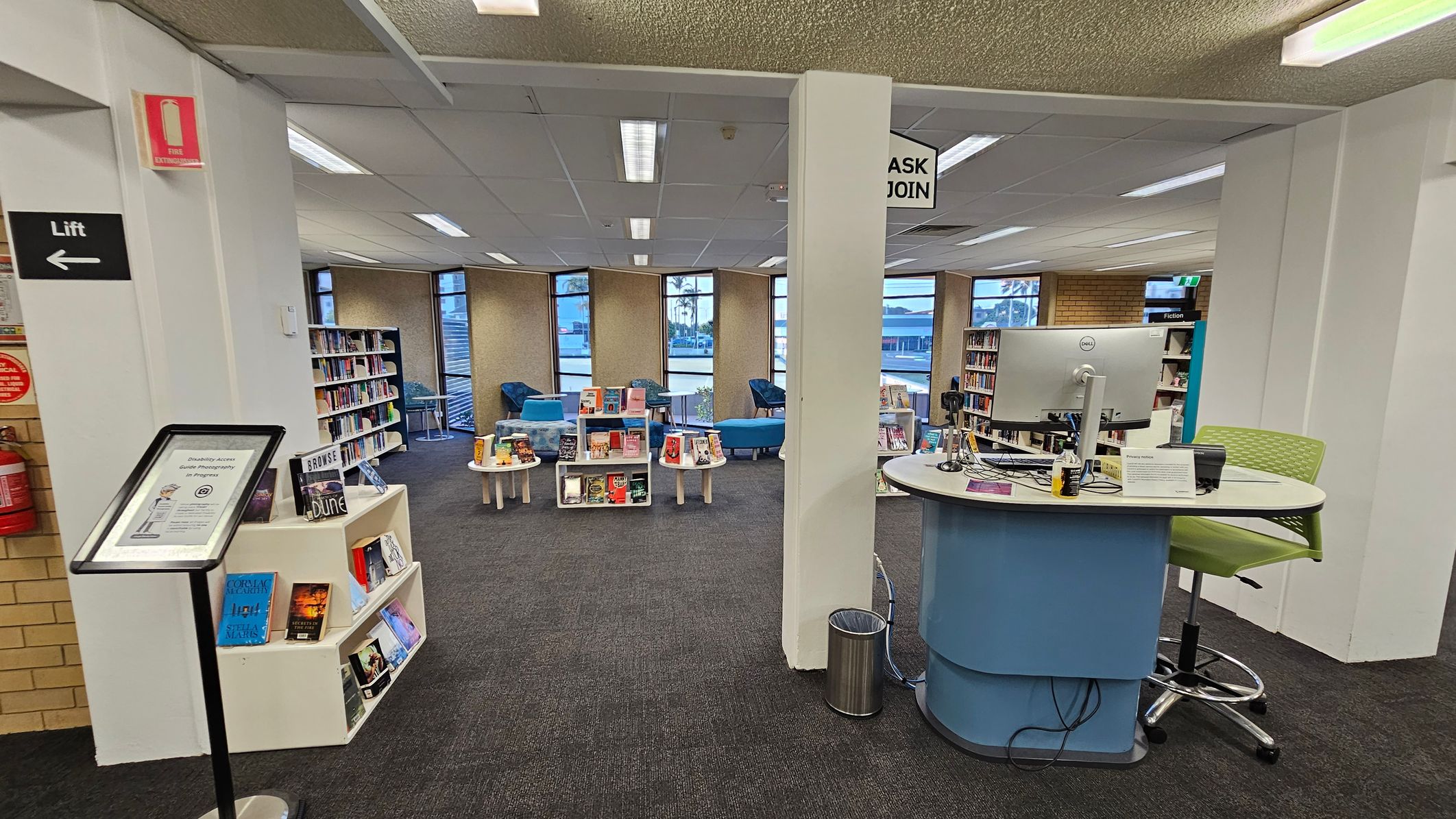 A library interior with a central desk on the right, featuring a computer and a green chair. In the middle, there are small tables and shelves displaying books. The left side has a sign for a lift and a fire extinguisher. Bookshelves line the walls, and there are blue chairs near the windows. The ceiling has fluorescent lights, and the floor is carpeted in dark grey.