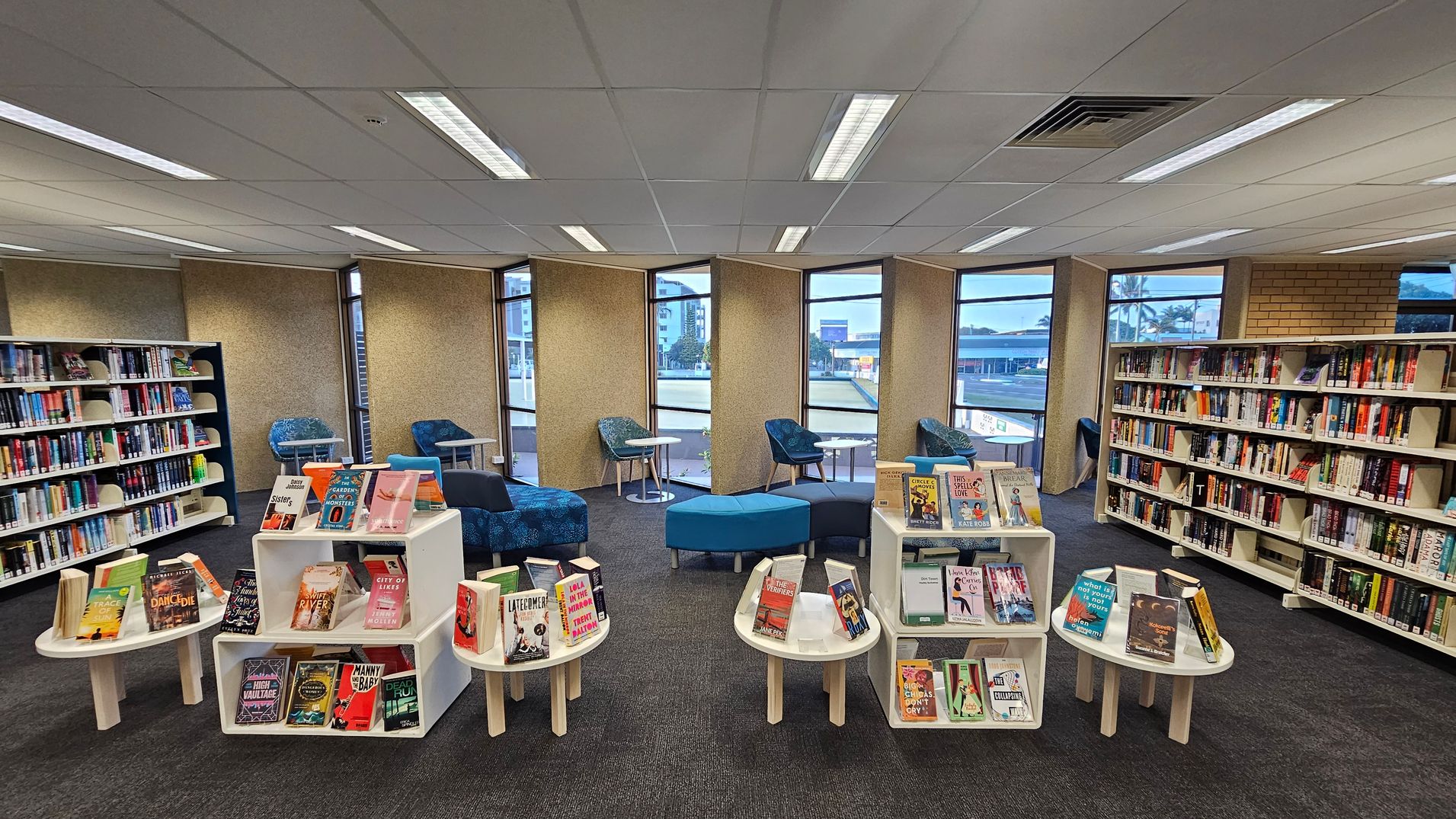 A library interior with bookshelves lining the left and right walls, filled with books. In the centre, there are white display tables showcasing various books. The background features large windows with views of buildings outside. Blue upholstered chairs and round tables are arranged near the windows. The floor is carpeted in dark grey, and the ceiling has white panels with fluorescent lighting.