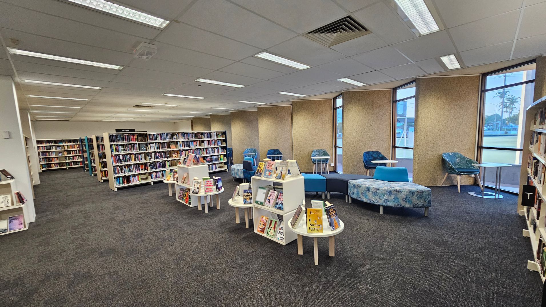 A library interior with bookshelves on the left filled with books. In the centre, there are white display stands showcasing various books. On the right, there are blue and patterned chairs with small round tables, and a curved seating area. The floor is carpeted in a mix of green and grey, and the ceiling has fluorescent lights. Large windows on the right wall allow natural light to enter.