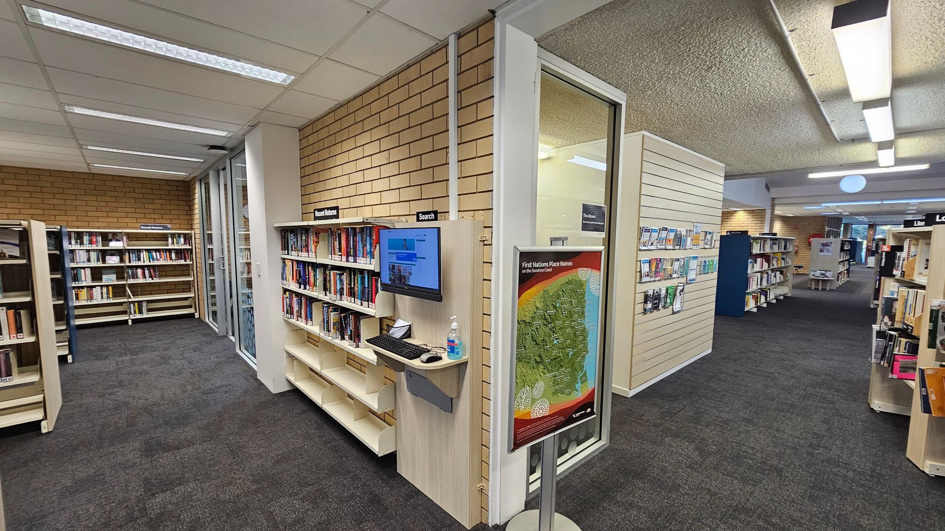 A library interior with bookshelves on the left and right. On the left, there are shelves labelled "Recent Returns" and a computer station for searching. A map titled "First Nations Place Names on the Sunshine Coast" is displayed on a stand. The floor is carpeted in dark grey, and the ceiling has fluorescent lights. In the background, more bookshelves and a seating area are visible.