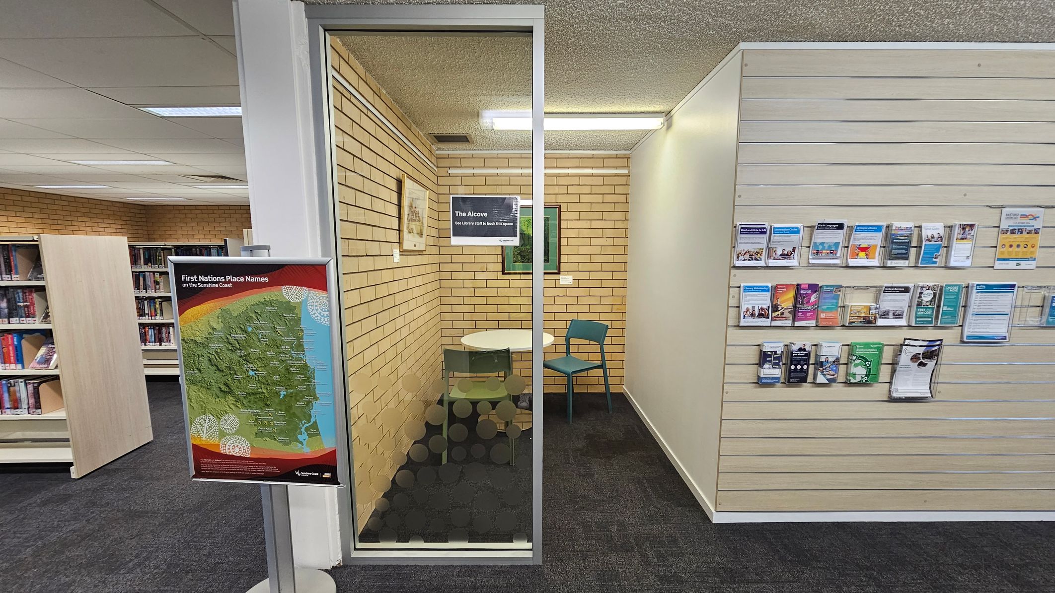 A library interior with a small glass-walled room labelled "The Alcove" in the centre. Inside, there is a round table and two chairs. The left side shows bookshelves filled with books. On the left, a stand displays a map titled "First Nations Place Names on the Sunshine Coast." The right side features a wall with various brochures and pamphlets neatly arranged. The floor is carpeted, and the ceiling has fluorescent lighting.