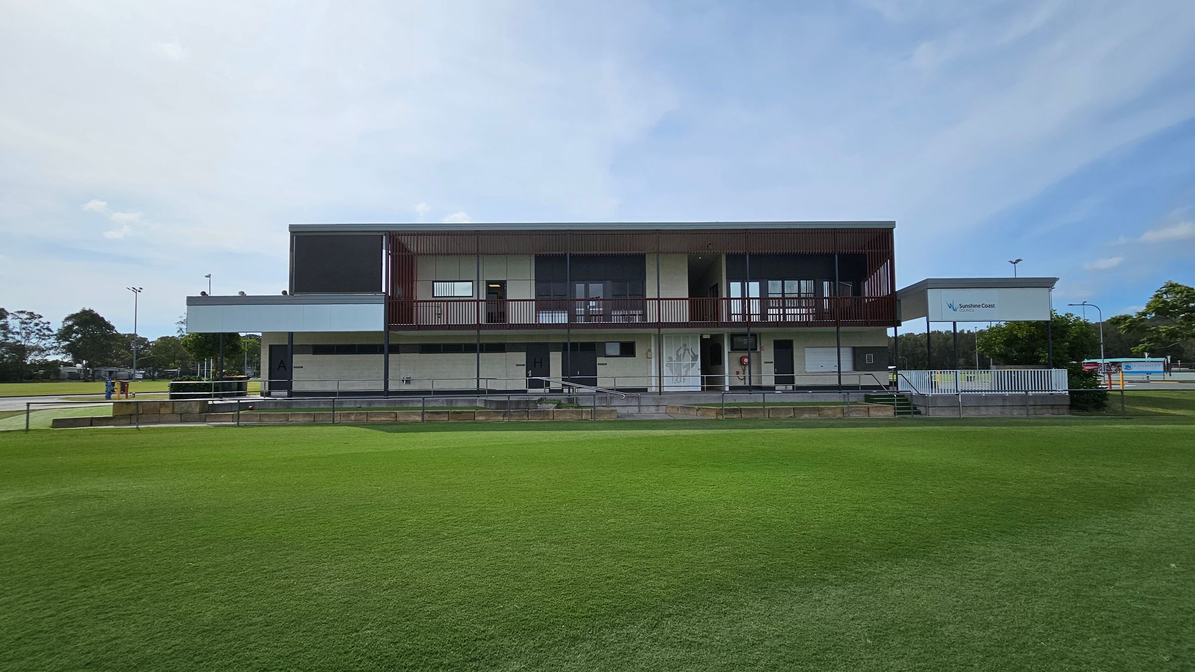 A modern, two-storey building with a flat roof and a mix of concrete and glass elements. The upper level features a balcony with railings, while the lower level has several doors and a ramp. The building is surrounded by a well-maintained grassy field. On the right, there's a sign with "Sunshine Coast" and some trees. In the background, there are more trees and a few light poles. A modern, two-storey building with a flat roof and a mix of concrete and glass elements. The upper level features a balcony with railings, while the lower level has several doors and a ramp. The building is surrounded by a well-maintained grassy field. On the right, there's a sign with "Sunshine Coast" and some trees. In the background, there are more trees and a few light poles.