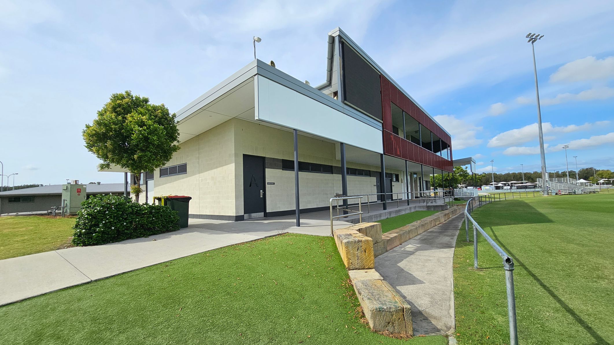 A modern sports facility with a two-storey building on the right, featuring a mix of cream and dark red panelling. A small tree and bushes are on the left near a concrete path. The foreground has a grassy area with a low stone wall and a metal railing. In the background, there are sports fields with tall floodlights and several white structures. The sky is clear with some clouds. A modern sports facility with a two-storey building on the right, featuring a mix of cream and dark red panelling. A small tree and bushes are on the left near a concrete path. The foreground has a grassy area with a low stone wall and a metal railing. In the background, there are sports fields with tall floodlights and several white structures. The sky is clear with some clouds.