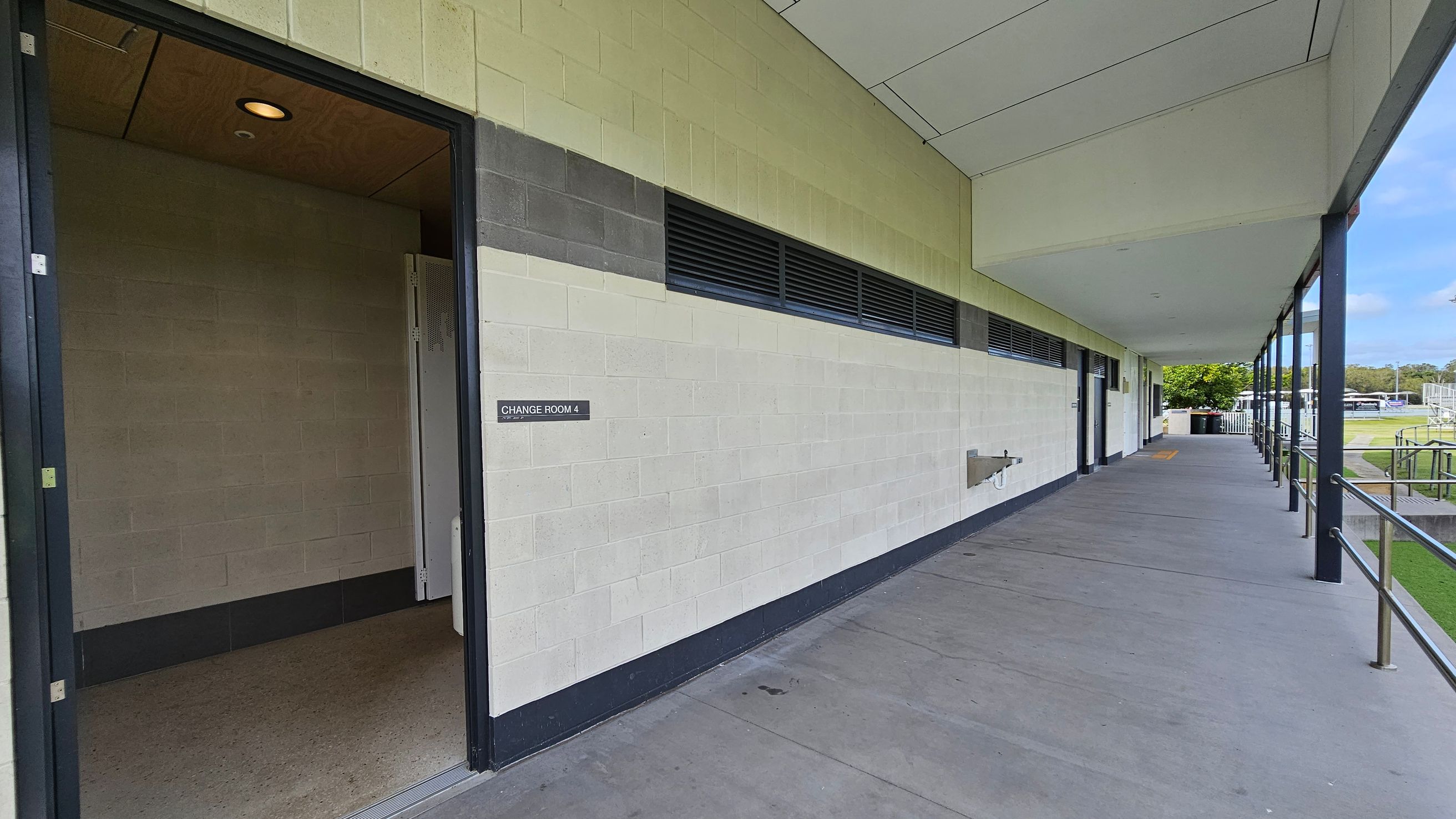 A corridor with a concrete floor and a ceiling with panels. On the left, there's an open door labelled "Change Room 4" leading into a room with a light-coloured floor. The wall is painted in light green and grey with ventilation panels. On the right, a railing runs along the edge of the corridor, and beyond it, there is a grassy area with trees and outdoor structures. The corridor extends into the distance with more doors and a water fountain visible. A corridor with a concrete floor and a ceiling with panels. On the left, there's an open door labelled "Change Room 4" leading into a room with a light-coloured floor. The wall is painted in light green and grey with ventilation panels. On the right, a railing runs along the edge of the corridor, and beyond it, there is a grassy area with trees and outdoor structures. The corridor extends into the distance with more doors and a water fountain visible.