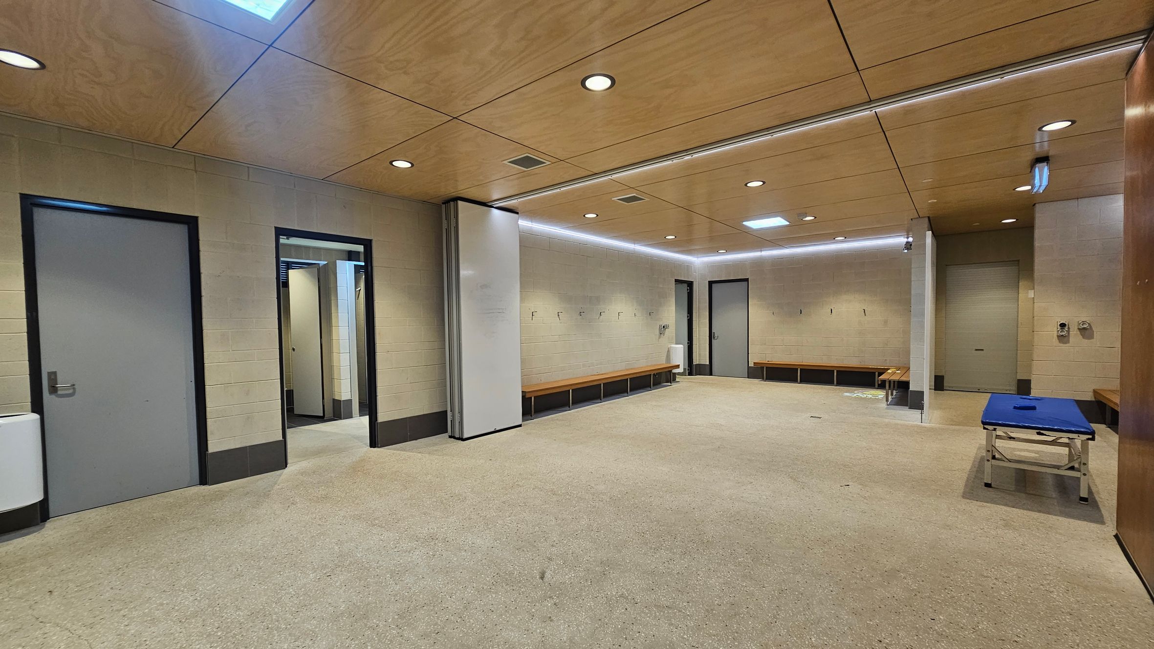 A spacious locker room with beige walls and a light brown ceiling featuring recessed lighting. On the left, there are two grey doors and a white wall-mounted unit. In the centre, a doorway leads to another area. Along the right wall, wooden benches are fixed, and a blue padded table is positioned nearby. The floor is covered with a light-coloured carpet. A spacious locker room with beige walls and a light brown ceiling featuring recessed lighting. On the left, there are two grey doors and a white wall-mounted unit. In the centre, a doorway leads to another area. Along the right wall, wooden benches are fixed, and a blue padded table is positioned nearby. The floor is covered with a light-coloured carpet.