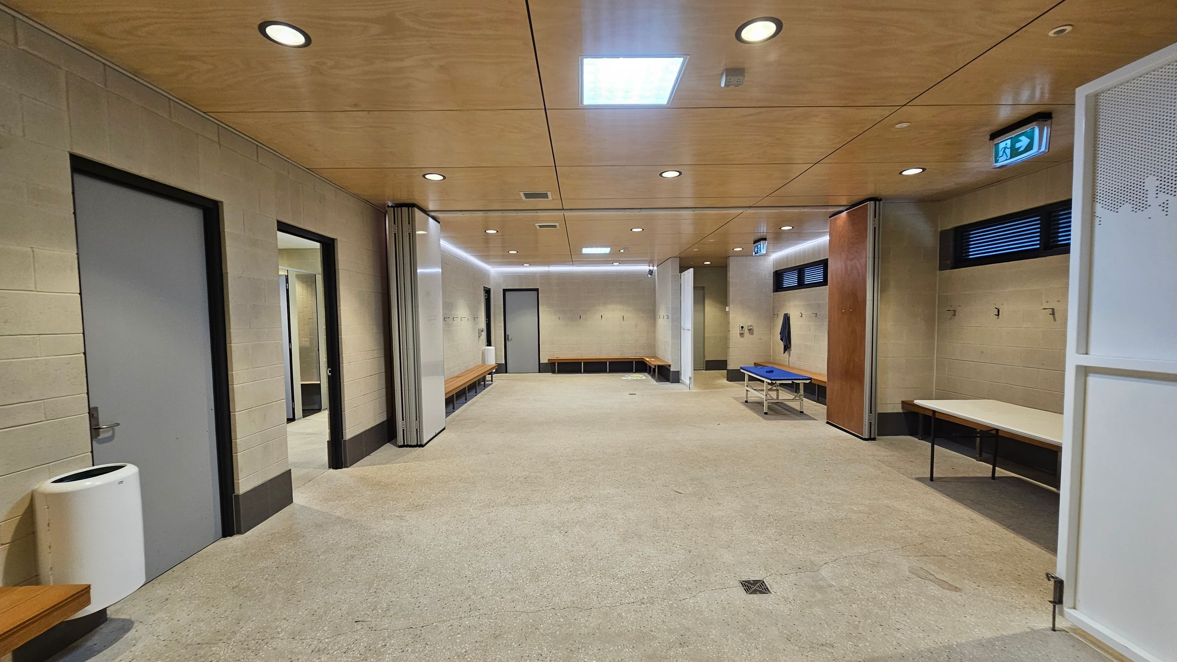 A spacious locker room with beige walls and a light-coloured ceiling featuring recessed lighting. On the left, there are grey doors and a white bin. Benches line the walls, with hooks above them. On the right, a blue bench and a table are visible. The floor is a speckled light grey. An emergency exit sign is above a door on the right. A spacious locker room with beige walls and a light-coloured ceiling featuring recessed lighting. On the left, there are grey doors and a white bin. Benches line the walls, with hooks above them. On the right, a blue bench and a table are visible. The floor is a speckled light grey. An emergency exit sign is above a door on the right.