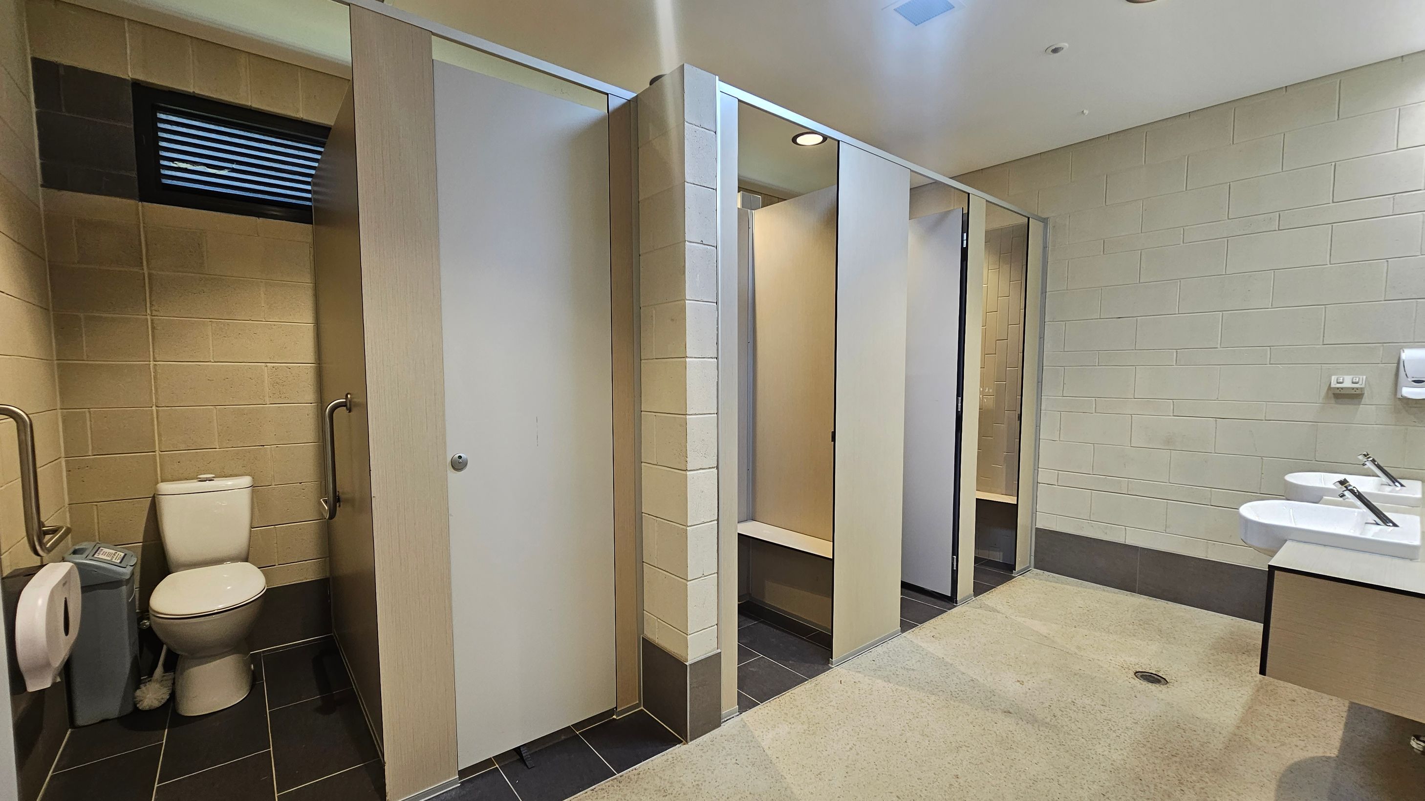 A public restroom with beige tiled walls and a light-coloured ceiling. On the left, there is a toilet cubicle with a white toilet, grab bars, a toilet paper dispenser, and a sanitary bin. The middle section has several closed cubicle doors. On the right, there are two white sinks with silver taps mounted on a countertop, and a soap dispenser on the wall. The floor is a mix of dark tiles and light-coloured flooring. A public restroom with beige tiled walls and a light-coloured ceiling. On the left, there is a toilet cubicle with a white toilet, grab bars, a toilet paper dispenser, and a sanitary bin. The middle section has several closed cubicle doors. On the right, there are two white sinks with silver taps mounted on a countertop, and a soap dispenser on the wall. The floor is a mix of dark tiles and light-coloured flooring.