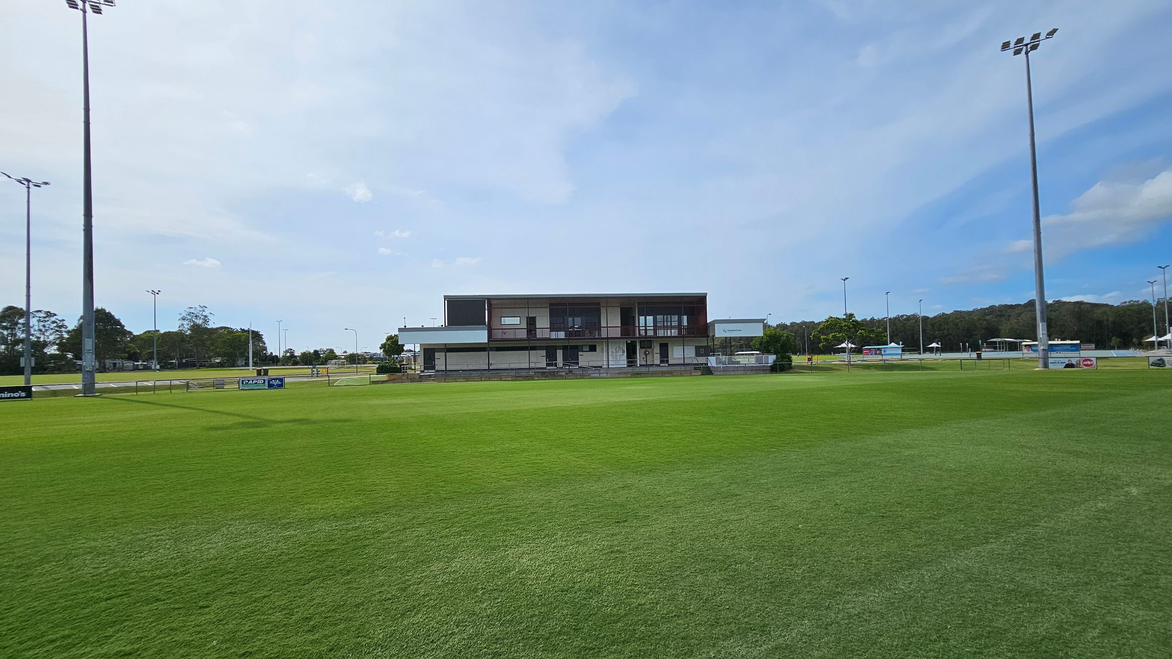 A sports field with well-maintained green grass in the foreground. In the background, there is a modern building with a flat roof and large windows, surrounded by a low wall. A tall floodlight pole stands prominently in the centre. To the left, there are additional structures and a few trees. On the right, a covered seating area with an orange panel is visible. The sky is partly cloudy. A sports field with well-maintained green grass in the foreground. In the background, there is a modern building with a flat roof and large windows, surrounded by a low wall. A tall floodlight pole stands prominently in the centre. To the left, there are additional structures and a few trees. On the right, a covered seating area with an orange panel is visible. The sky is partly cloudy.