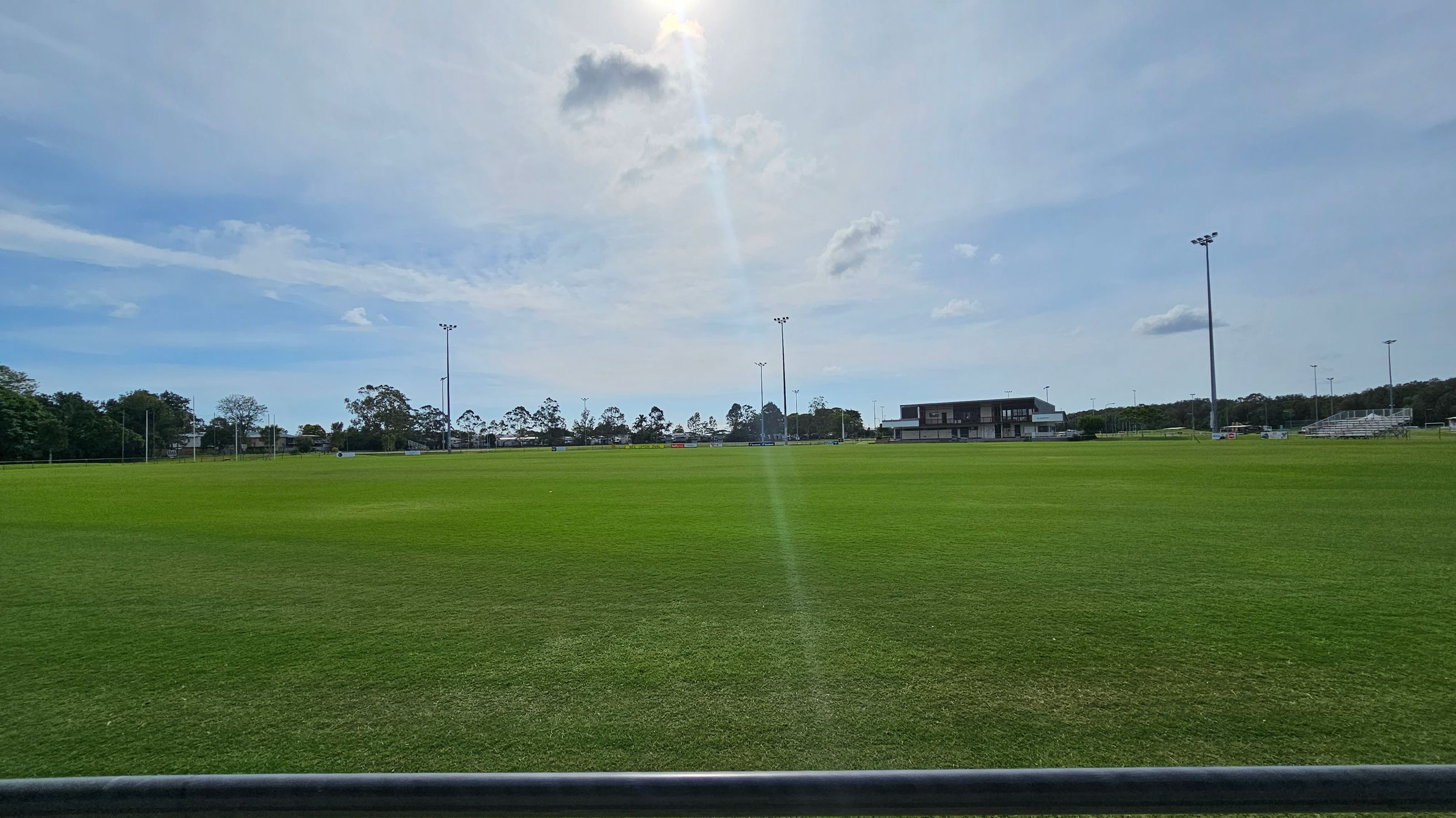 A large, open grassy field with a few patches of brown. In the background, there are several tall floodlights and a building to the right. Trees line the horizon, and there are some small structures or equipment scattered across the field. The sky is mostly clear with a few clouds. A large, open grassy field with a few patches of brown. In the background, there are several tall floodlights and a building to the right. Trees line the horizon, and there are some small structures or equipment scattered across the field. The sky is mostly clear with a few clouds.