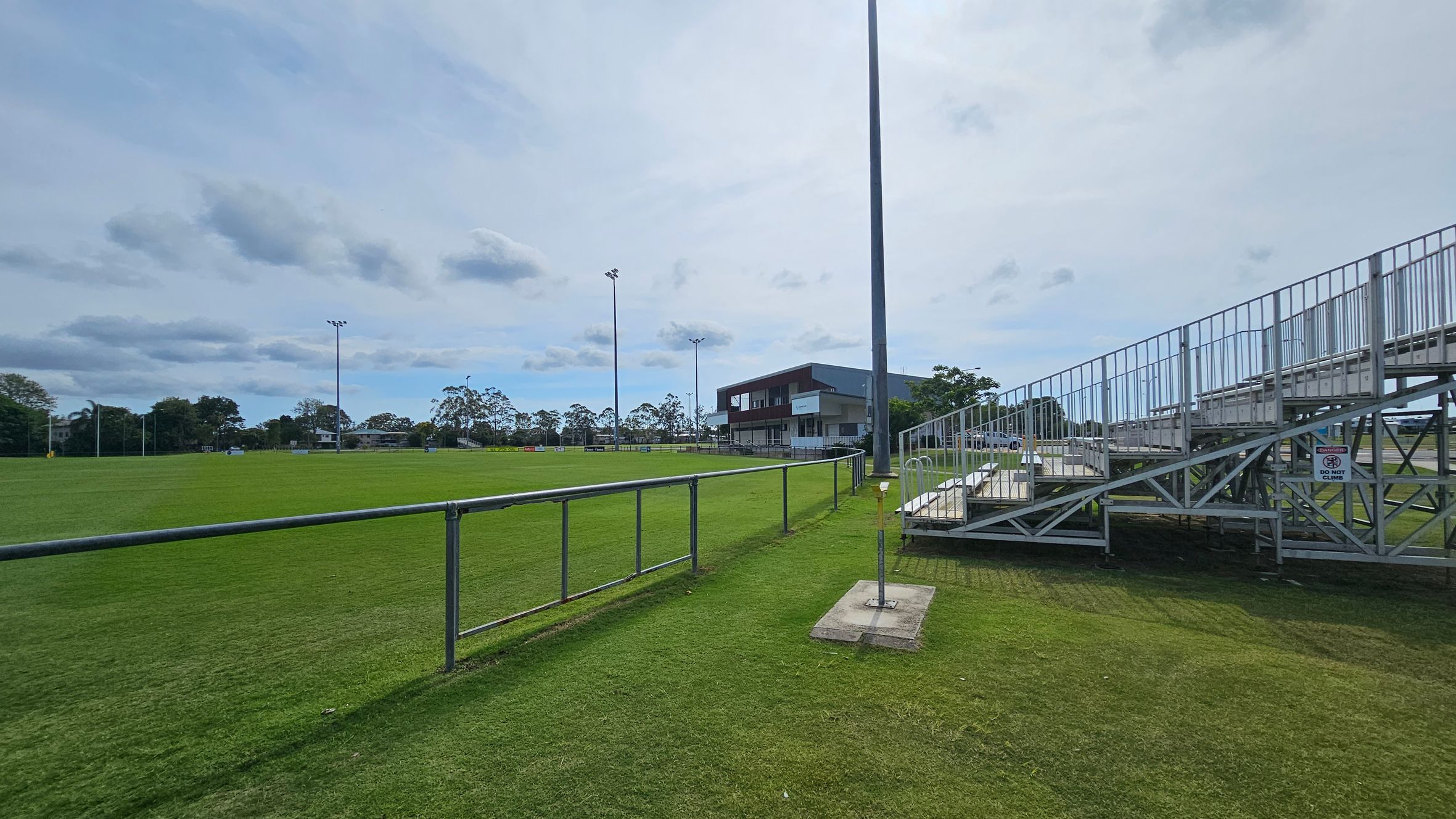 A sports field with a well-maintained grass surface. On the right, there are metal bleachers with a sign that reads "DO NOT CLIMB." In the centre, a tall floodlight pole stands near a building with a balcony. The background features trees and additional floodlights. A metal railing runs along the left side of the field. A sports field with a well-maintained grass surface. On the right, there are metal bleachers with a sign that reads "DO NOT CLIMB." In the centre, a tall floodlight pole stands near a building with a balcony. The background features trees and additional floodlights. A metal railing runs along the left side of the field.
