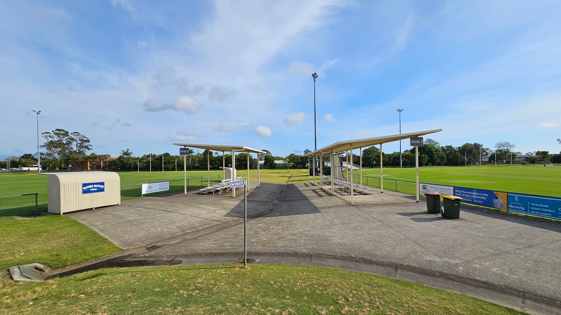 A sports field with two covered seating areas in the centre, labelled "Oval 1" and "Oval 2". The ground is a mix of concrete and grass. A small structure on the left has a sign reading "Panther Match Day Quarters". A signpost indicates the direction to toilets. Two bins are near the seating on the right. The background features a grassy field with goalposts and trees. Advertising banners line the fence on the right. A sports field with two covered seating areas in the centre, labelled "Oval 1" and "Oval 2". The ground is a mix of concrete and grass. A small structure on the left has a sign reading "Panther Match Day Quarters". A signpost indicates the direction to toilets. Two bins are near the seating on the right. The background features a grassy field with goalposts and trees. Advertising banners line the fence on the right.