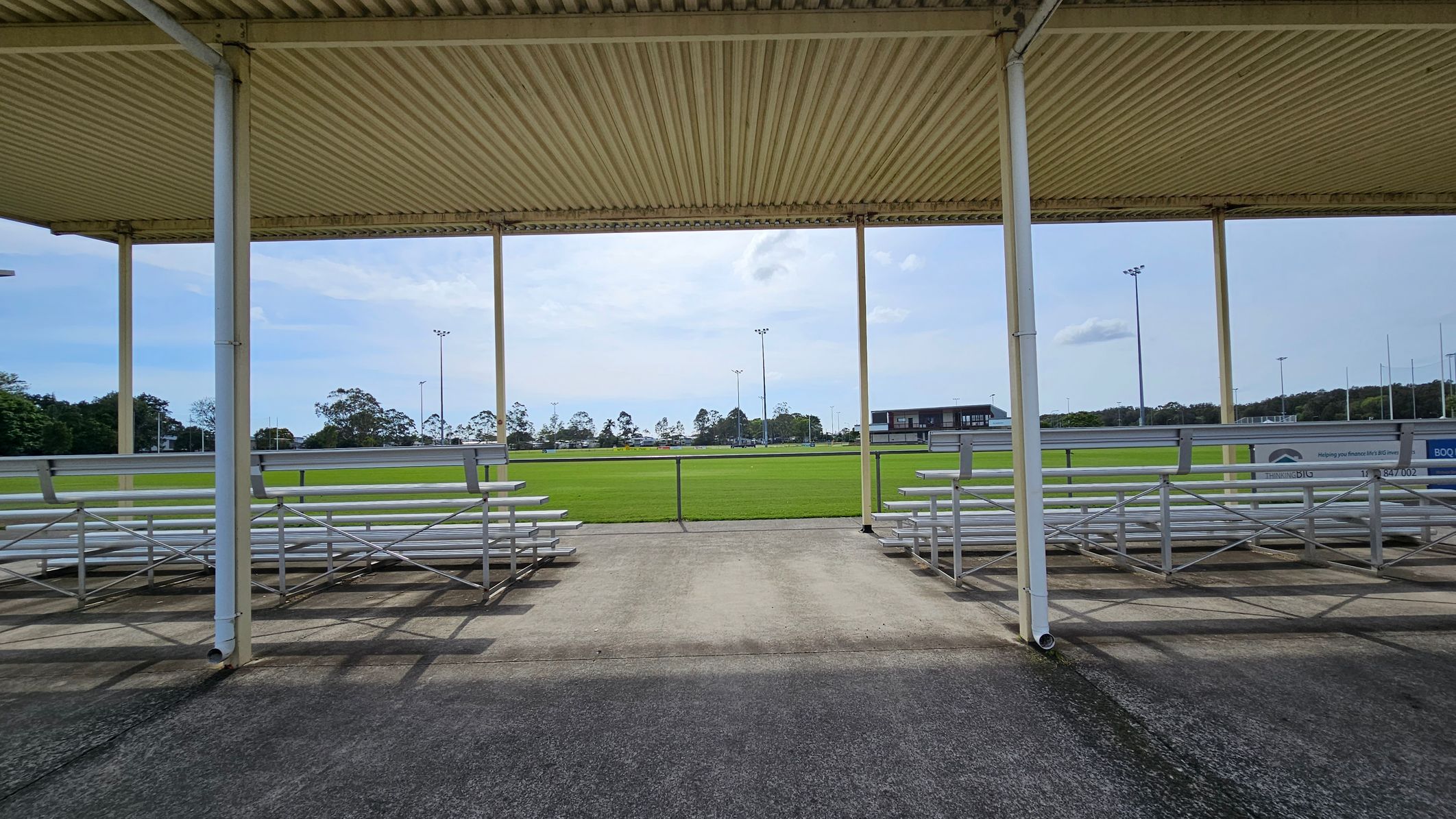 A sports field with a green grass pitch in the background, surrounded by tall floodlights. In the foreground, there are metal bleachers on both the left and right sides, under a corrugated metal roof supported by white poles. The ground is paved with concrete, and there are trees and a building visible in the distance. A sports field with a green grass pitch in the background, surrounded by tall floodlights. In the foreground, there are metal bleachers on both the left and right sides, under a corrugated metal roof supported by white poles. The ground is paved with concrete, and there are trees and a building visible in the distance.