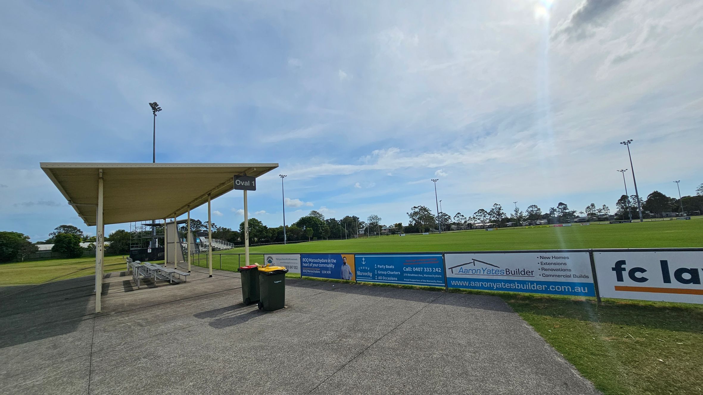 A sports field with a green grass pitch is in the centre, surrounded by a low fence with advertising banners. On the left, there's a covered seating area with benches and a sign reading "Oval 1". Two rubbish bins are placed nearby on a paved path. Tall floodlights are visible around the field, and trees line the background. The sky is overcast. A sports field with a green grass pitch is in the centre, surrounded by a low fence with advertising banners. On the left, there's a covered seating area with benches and a sign reading "Oval 1". Two rubbish bins are placed nearby on a paved path. Tall floodlights are visible around the field, and trees line the background. The sky is overcast.