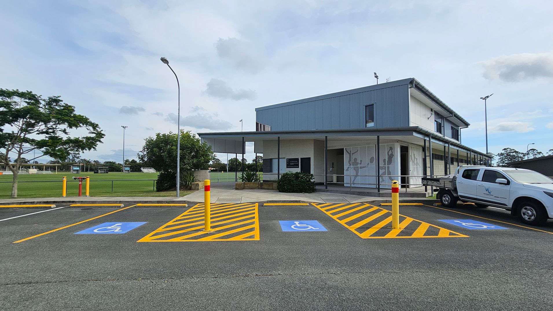 A modern building with a grey exterior is on the right, featuring large windows and a covered entrance. In front, there are marked disabled parking spaces with blue symbols and yellow lines. A white utility vehicle is parked in one of the spaces. On the left, there is a grassy field with a few trees and a tall streetlamp. The background includes more greenery and sports fields. The ground is paved, and there are yellow bollards near the parking area. A modern building with a grey exterior is on the right, featuring large windows and a covered entrance. In front, there are marked disabled parking spaces with blue symbols and yellow lines. A white utility vehicle is parked in one of the spaces. On the left, there is a grassy field with a few trees and a tall streetlamp. The background includes more greenery and sports fields. The ground is paved, and there are yellow bollards near the parking area.