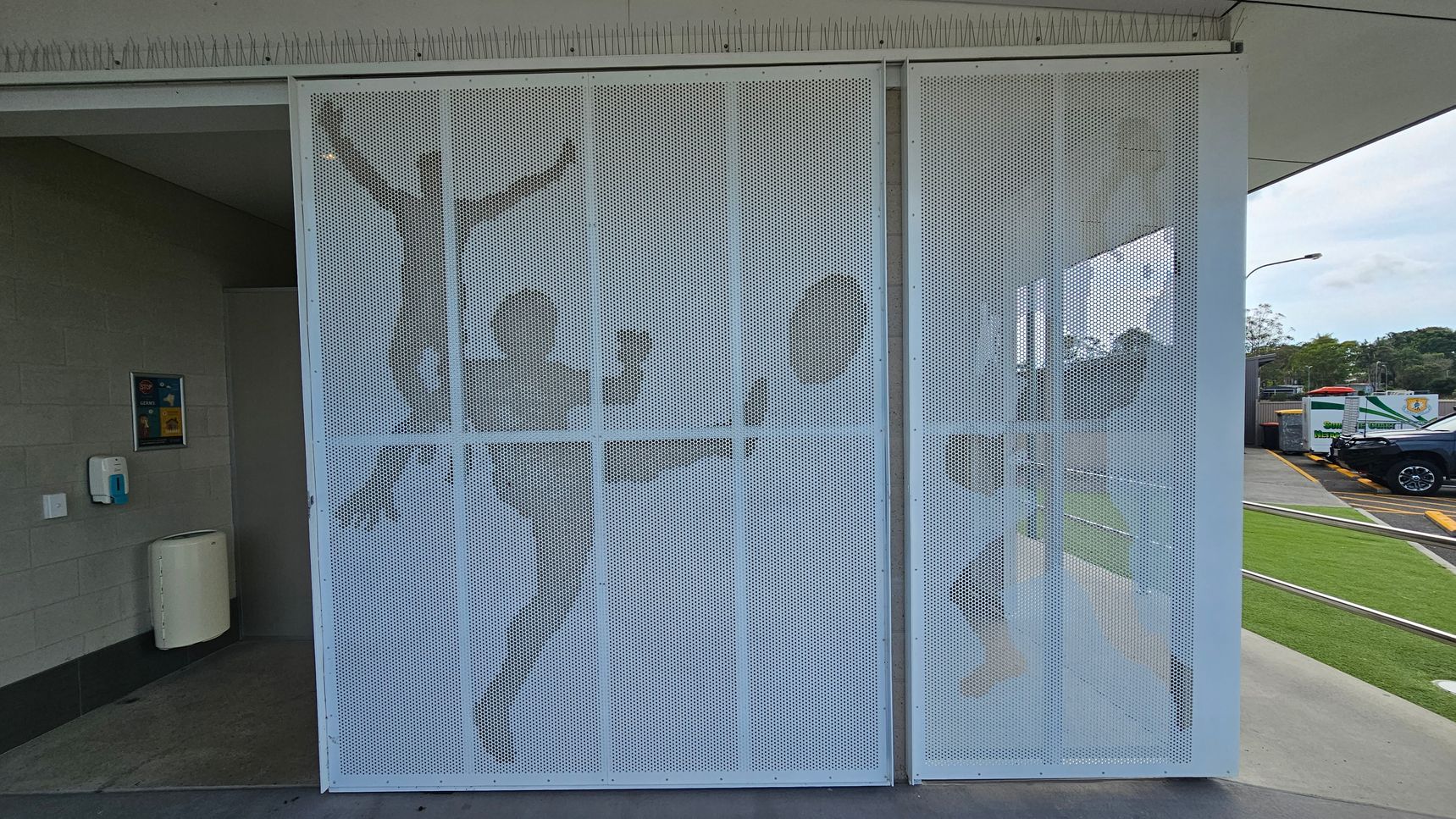 A perforated metal screen with silhouettes of people engaged in athletic activities, such as kicking a ball, is positioned in the centre. On the left, there's a wall with a hand sanitiser dispenser and a waste bin. The right side shows a parking area with a black vehicle and some greenery. The ceiling has a row of anti-bird spikes. A perforated metal screen with silhouettes of people engaged in athletic activities, such as kicking a ball, is positioned in the centre. On the left, there's a wall with a hand sanitiser dispenser and a waste bin. The right side shows a parking area with a black vehicle and some greenery. The ceiling has a row of anti-bird spikes.