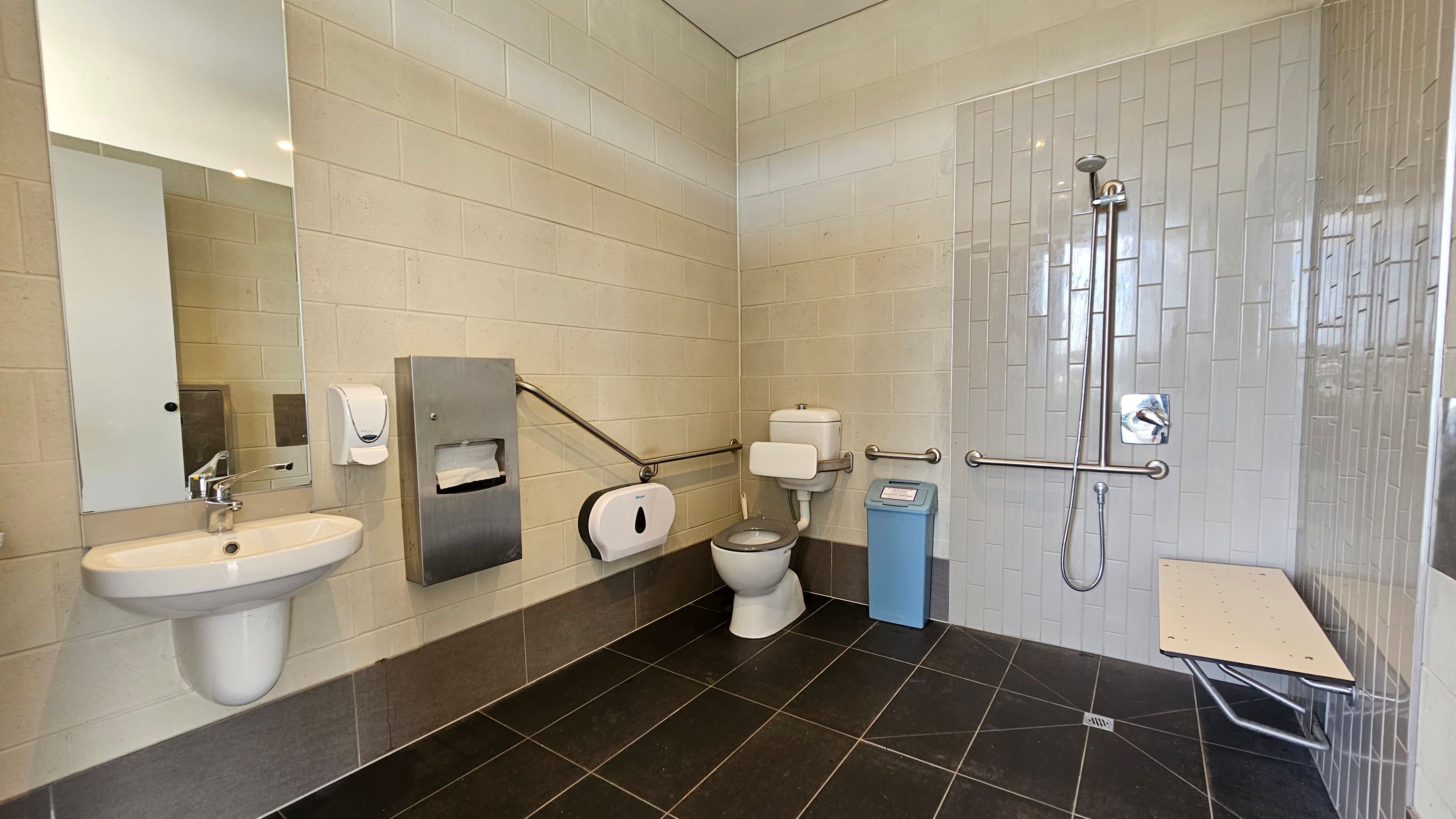 Accessible bathroom with a white sink and mirror on the left. A soap dispenser and paper towel holder are mounted on the wall. In the centre, a toilet with support rails and a blue bin nearby. On the right, a shower area with a handheld showerhead and grab bars, alongside a foldable bench. The walls are tiled in light colours, and the floor has dark tiles. Accessible bathroom with a white sink and mirror on the left. A soap dispenser and paper towel holder are mounted on the wall. In the centre, a toilet with support rails and a blue bin nearby. On the right, a shower area with a handheld showerhead and grab bars, alongside a foldable bench. The walls are tiled in light colours, and the floor has dark tiles.