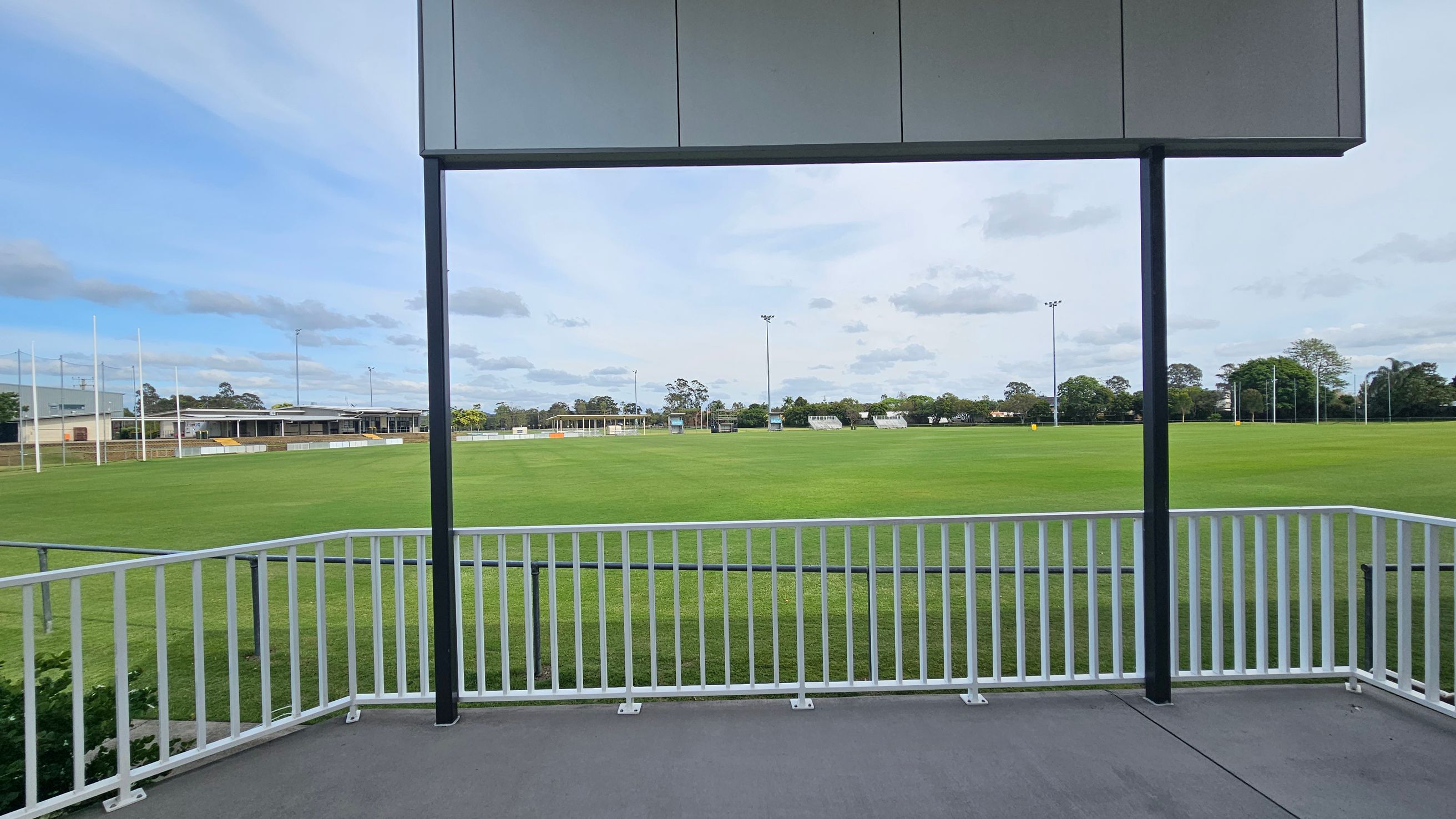 A sports field with a well-maintained green lawn is visible, surrounded by a white railing in the foreground. In the background, there are goalposts and a building with a flat roof on the left. Trees and additional goalposts are scattered along the horizon. A metal structure with a roof is partially visible at the top of the image. A sports field with a well-maintained green lawn is visible, surrounded by a white railing in the foreground. In the background, there are goalposts and a building with a flat roof on the left. Trees and additional goalposts are scattered along the horizon. A metal structure with a roof is partially visible at the top of the image.
