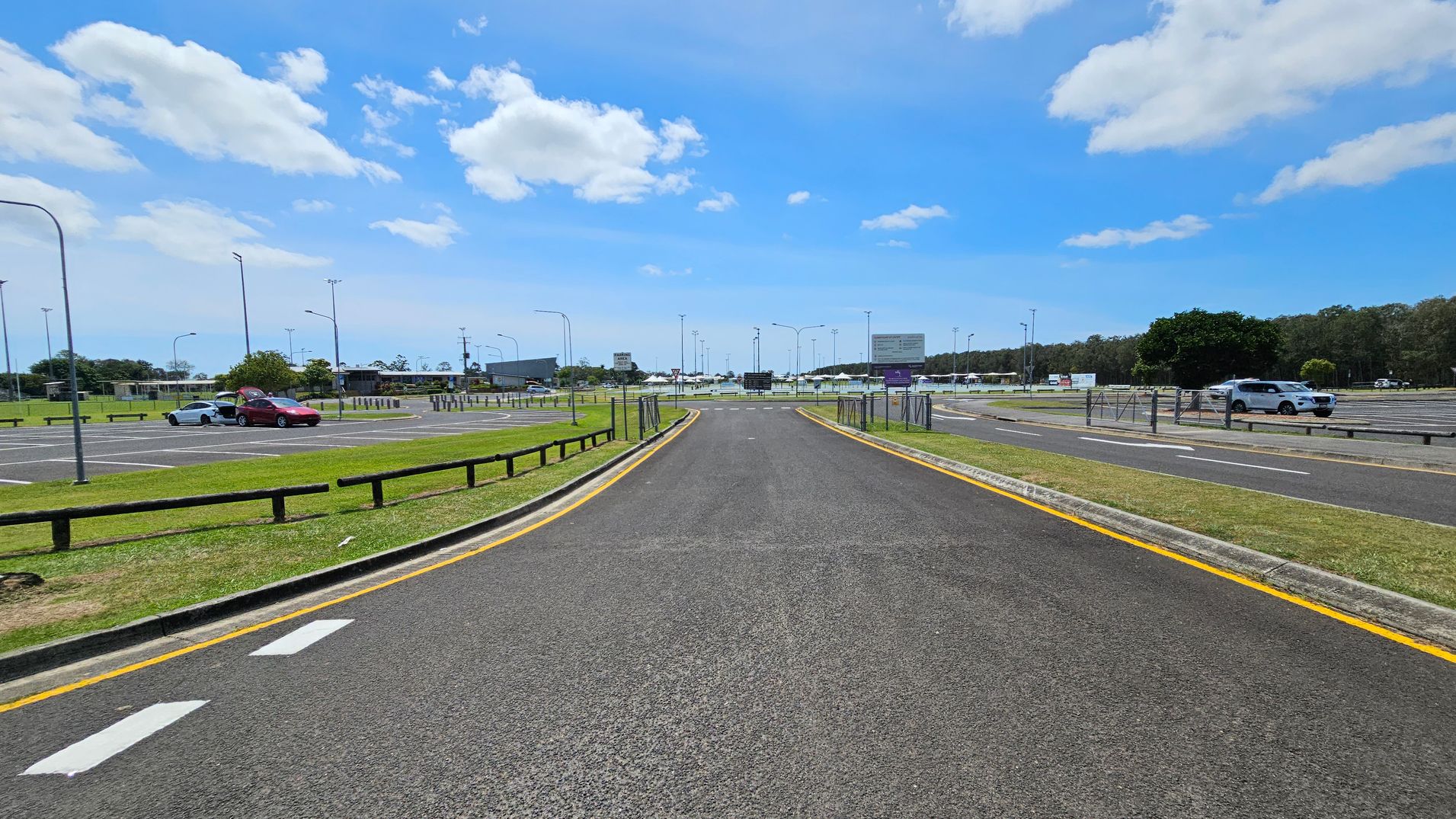 A road curves to the left with a yellow line marking its edge. On the right, a blue sign reads "Maroochydore Multi Sports Complex." Beyond the road, there are parking spaces with a few cars and a grassy area with trees in the background. Several streetlights and signs are visible along the road. The sky is clear with a few clouds. A road curves to the left with a yellow line marking its edge. On the right, a blue sign reads "Maroochydore Multi Sports Complex." Beyond the road, there are parking spaces with a few cars and a grassy area with trees in the background. Several streetlights and signs are visible along the road. The sky is clear with a few clouds.