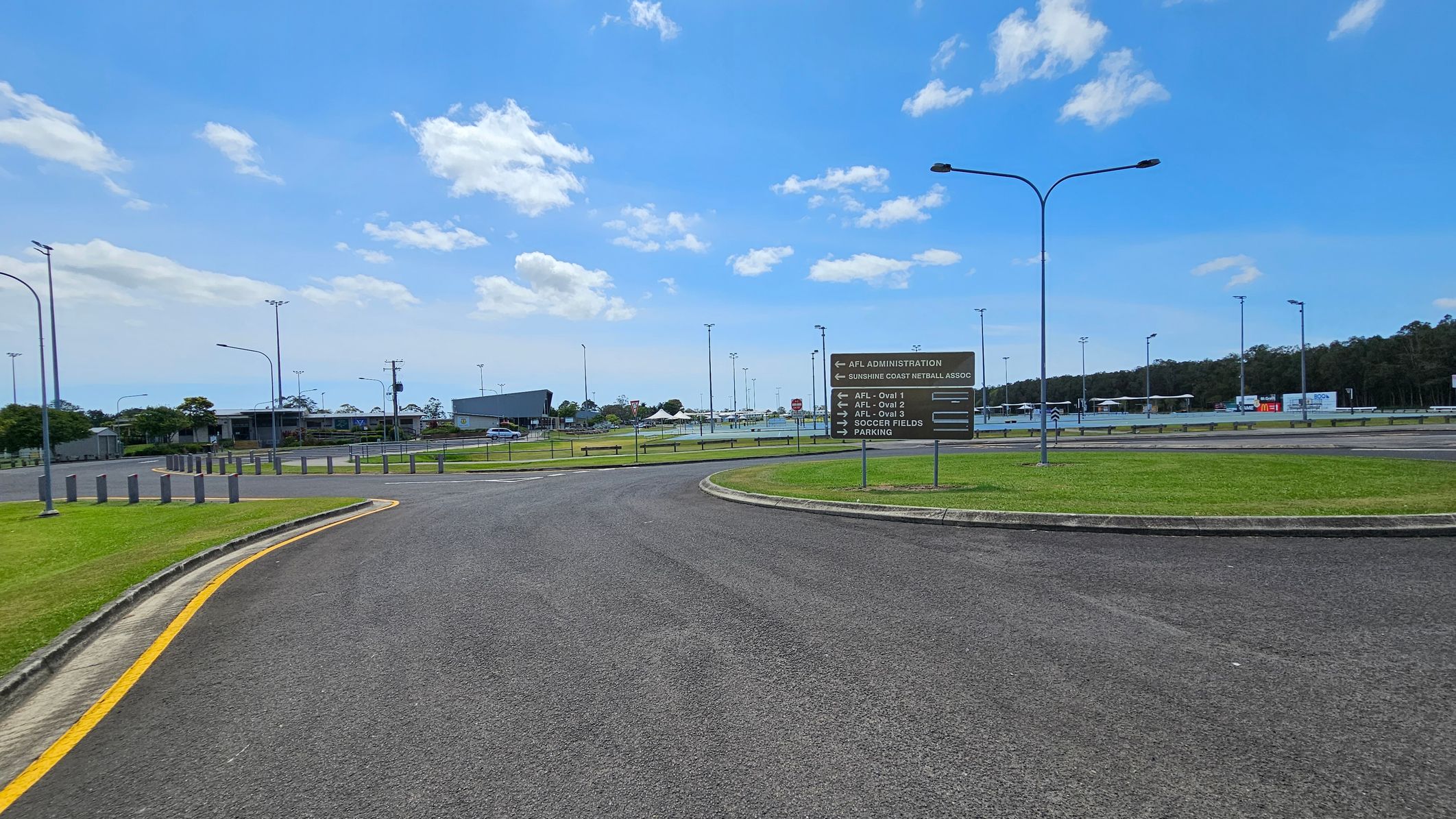 A paved road curves to the left, bordered by grass with a yellow line marking the edge. On the right, a sign lists directions to various sports facilities, including AFL ovals and soccer fields. In the background, there are several buildings, light poles, and a parking area. Trees line the horizon on the right side. The sky is partly cloudy. A paved road curves to the left, bordered by grass with a yellow line marking the edge. On the right, a sign lists directions to various sports facilities, including AFL ovals and soccer fields. In the background, there are several buildings, light poles, and a parking area. Trees line the horizon on the right side. The sky is partly cloudy.