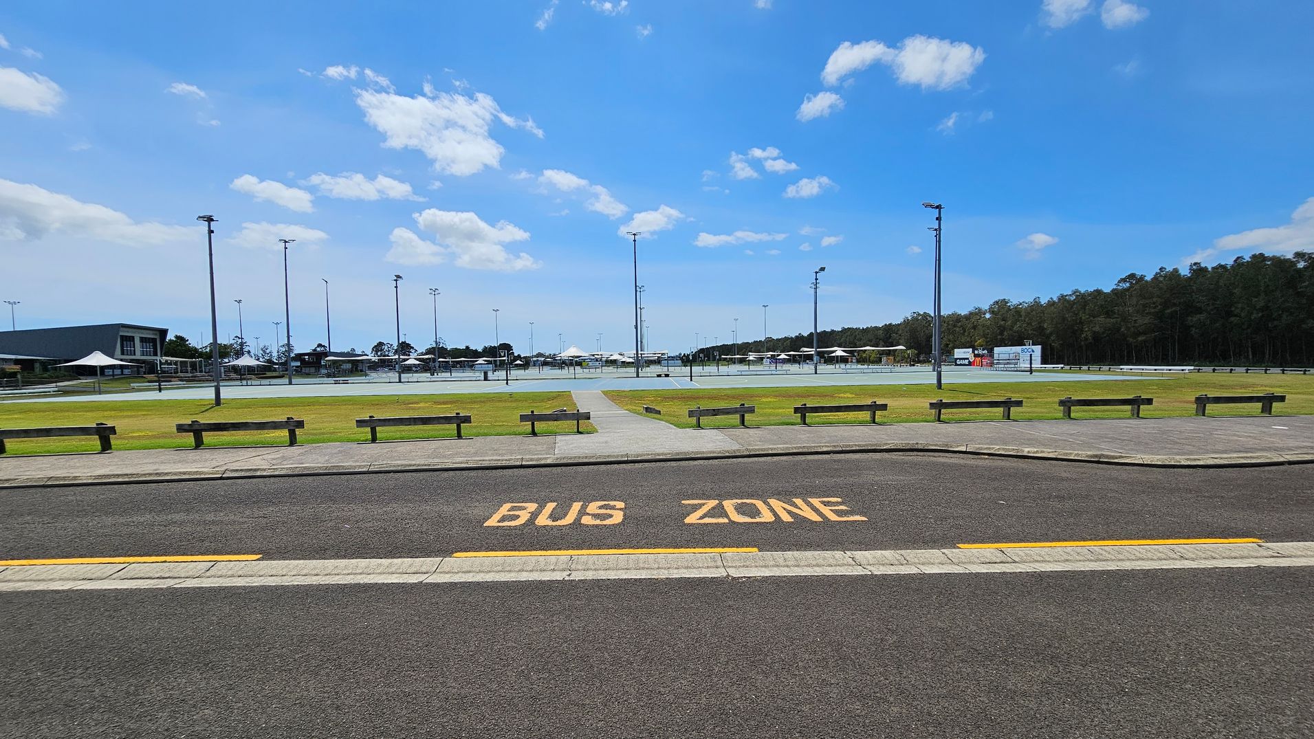 A paved road with "BUS ZONE" painted in yellow is in the foreground. Beyond the road, there is a grassy area with several wooden benches and a path leading to a sports complex. The complex features multiple light poles and a large building on the left. In the background, there are more structures and a line of trees on the right. A paved road with "BUS ZONE" painted in yellow is in the foreground. Beyond the road, there is a grassy area with several wooden benches and a path leading to a sports complex. The complex features multiple light poles and a large building on the left. In the background, there are more structures and a line of trees on the right.