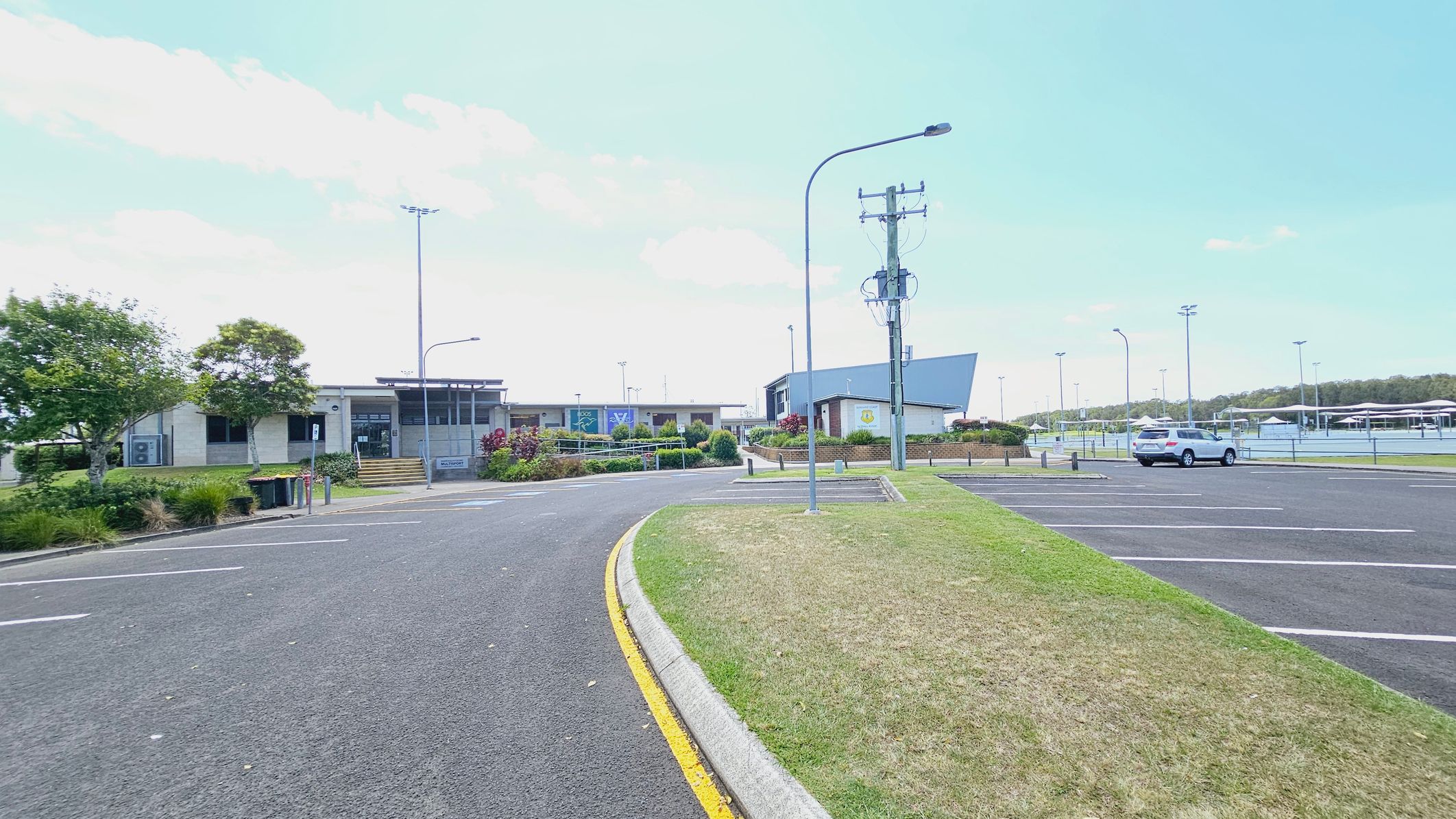 A car park with a single white vehicle on the right. The foreground features a curved road with a yellow line and adjacent grass. In the background, there are buildings with modern architecture, surrounded by landscaped greenery. Several tall streetlights and a utility pole are visible. Tennis courts with fences are on the far right. A car park with a single white vehicle on the right. The foreground features a curved road with a yellow line and adjacent grass. In the background, there are buildings with modern architecture, surrounded by landscaped greenery. Several tall streetlights and a utility pole are visible. Tennis courts with fences are on the far right.