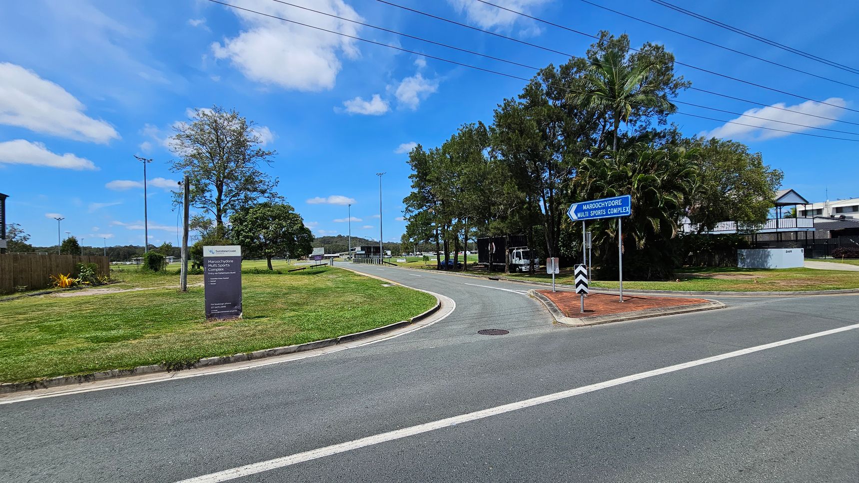 A road curves to the right, bordered by green grass and trees. On the left, a sign reads "Maroochydore Multi Sports Complex" with additional details. A blue directional sign points towards the complex. Trees and a few buildings are visible in the background. Overhead, power lines stretch across the scene. The road surface is grey with white markings. A road curves to the right, bordered by green grass and trees. On the left, a sign reads "Maroochydore Multi Sports Complex" with additional details. A blue directional sign points towards the complex. Trees and a few buildings are visible in the background. Overhead, power lines stretch across the scene. The road surface is grey with white markings.