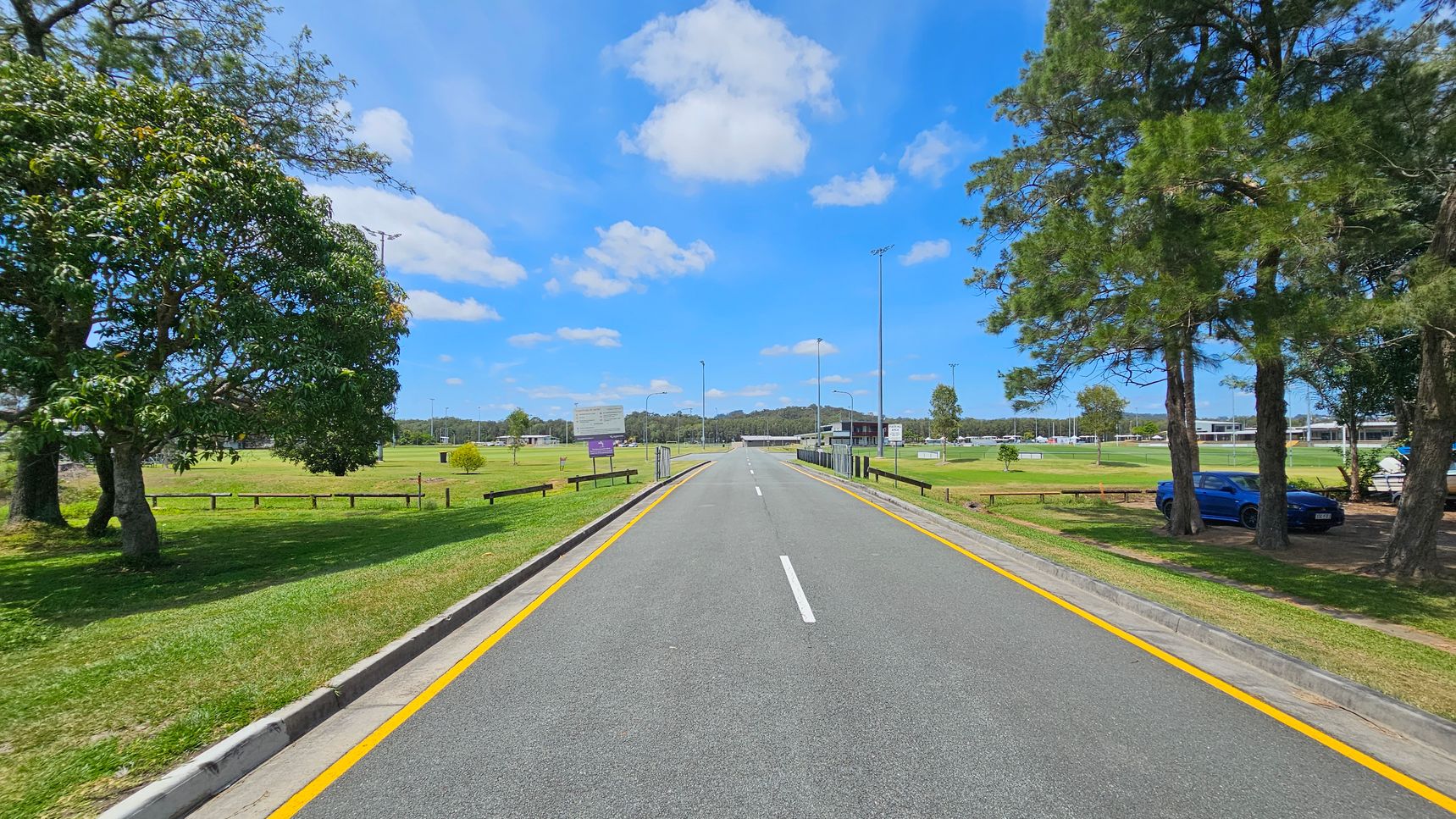 A straight road with yellow lines runs through the centre, flanked by grassy areas. On the left, there are several large trees and a wooden fence. On the right, more trees are visible, with a blue car parked under them. In the background, there are sports fields with goalposts and a few buildings. The sky is clear with scattered clouds. A straight road with yellow lines runs through the centre, flanked by grassy areas. On the left, there are several large trees and a wooden fence. On the right, more trees are visible, with a blue car parked under them. In the background, there are sports fields with goalposts and a few buildings. The sky is clear with scattered clouds.
