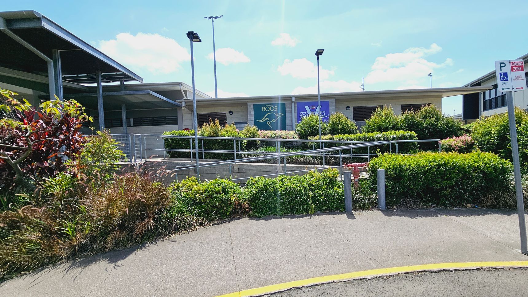 A building with a flat roof is in the centre, featuring a covered entrance with steps leading up to it. In front, there are several marked disabled parking spaces on an asphalt surface. To the left, a grassy area with a tree and two bins is visible. On the right, there are bushes and a signpost. The background shows a sports field with goalposts and more greenery. A building with a flat roof is in the centre, featuring a covered entrance with steps leading up to it. In front, there are several marked disabled parking spaces on an asphalt surface. To the left, a grassy area with a tree and two bins is visible. On the right, there are bushes and a signpost. The background shows a sports field with goalposts and more greenery.