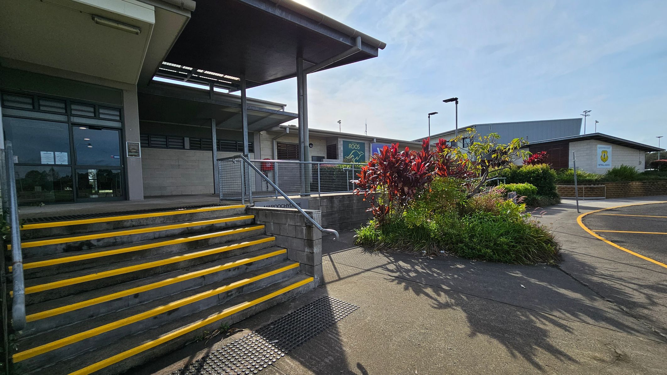 Concrete steps with yellow edges lead up to a building entrance on the left, featuring large glass windows and a metal railing. In the centre, a garden area with red and green plants is bordered by a concrete path. On the right, a curved road with yellow markings is visible. The building has a flat roof and a sign with a logo on the wall. The area is well-lit, and there are street lamps in the background. Concrete steps with yellow edges lead up to a building entrance on the left, featuring large glass windows and a metal railing. In the centre, a garden area with red and green plants is bordered by a concrete path. On the right, a curved road with yellow markings is visible. The building has a flat roof and a sign with a logo on the wall. The area is well-lit, and there are street lamps in the background.