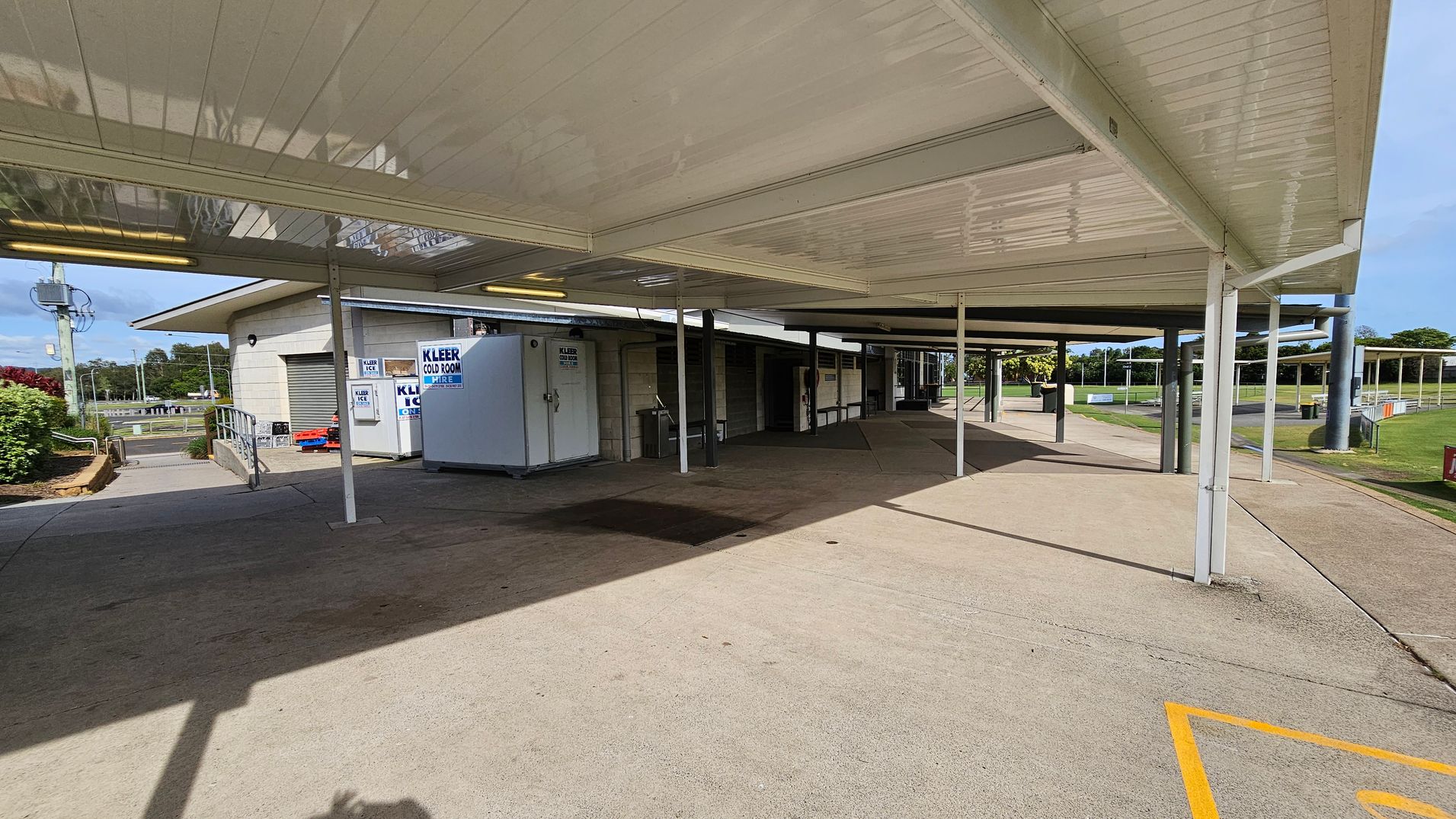 A covered walkway with a white ceiling and supporting poles. On the left, there's a building with a ramp leading to a door, and signs for "Kleer Cold Room Hire" and "Kleer Ice." A large white cold room unit is visible. The ground is concrete, and there are shadows from the structure. In the background, there is a grassy area with trees and a road. A covered walkway with a white ceiling and supporting poles. On the left, there's a building with a ramp leading to a door, and signs for "Kleer Cold Room Hire" and "Kleer Ice." A large white cold room unit is visible. The ground is concrete, and there are shadows from the structure. In the background, there is a grassy area with trees and a road.