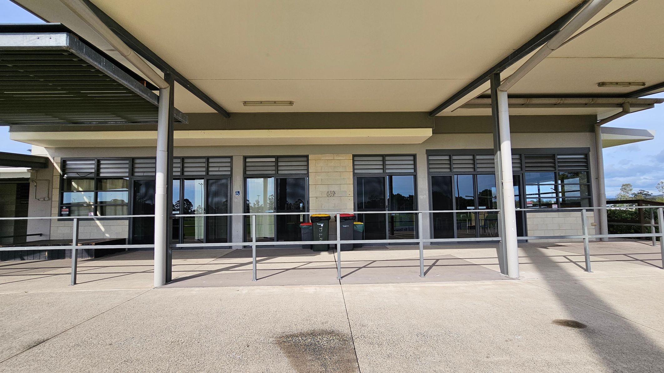 A building with a flat roof supported by metal columns. The facade features large glass windows with louvres above them. In front of the windows, there is a metal railing and several bins with coloured lids. The ground is paved with concrete, and there is a shadow cast by the roof. The background shows some greenery and a partly cloudy sky. A building with a flat roof supported by metal columns. The facade features large glass windows with louvres above them. In front of the windows, there is a metal railing and several bins with coloured lids. The ground is paved with concrete, and there is a shadow cast by the roof. The background shows some greenery and a partly cloudy sky.