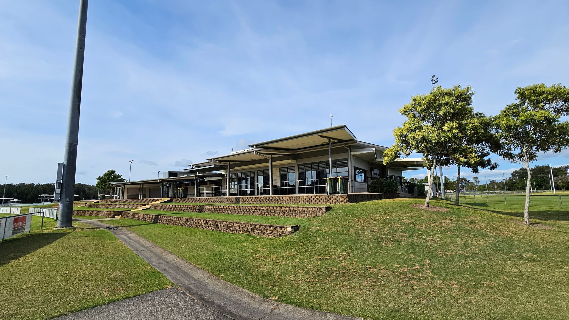 A modern building with a flat roof and large windows is situated on a grassy hill. In the foreground, there are tiered brick steps leading up to the building. To the right, several trees with green foliage are visible. On the left, a tall light pole stands near a pathway that runs alongside the grass. In the background, there are sports fields with goalposts and more trees. A modern building with a flat roof and large windows is situated on a grassy hill. In the foreground, there are tiered brick steps leading up to the building. To the right, several trees with green foliage are visible. On the left, a tall light pole stands near a pathway that runs alongside the grass. In the background, there are sports fields with goalposts and more trees.