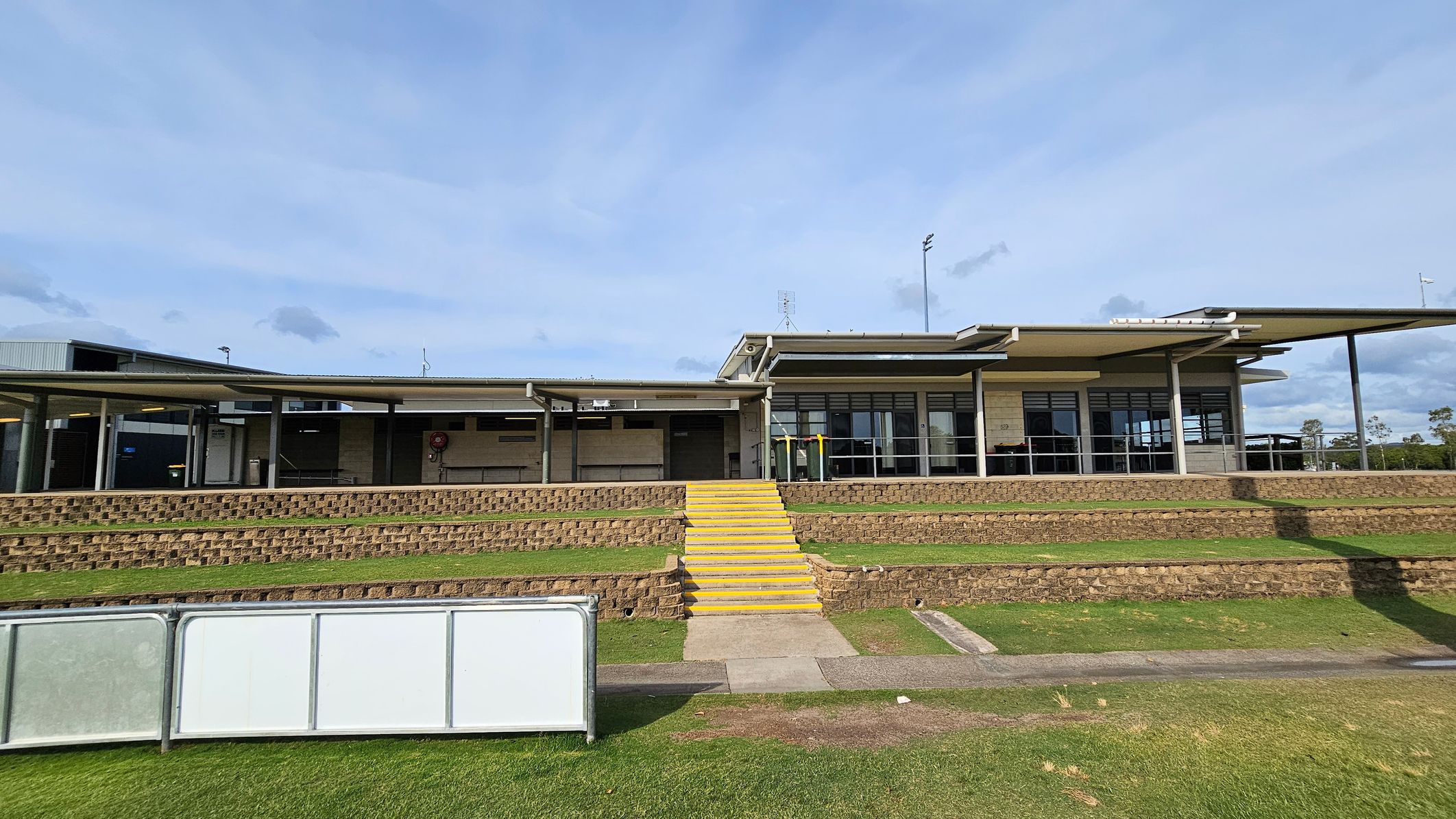 A modern building with a flat roof and large windows is in the centre. It has a series of yellow steps leading up to it, flanked by tiered brick retaining walls. The foreground features a grassy area with a concrete path and a white barrier. The building has a covered walkway supported by columns, and there are some trees visible in the background. A modern building with a flat roof and large windows is in the centre. It has a series of yellow steps leading up to it, flanked by tiered brick retaining walls. The foreground features a grassy area with a concrete path and a white barrier. The building has a covered walkway supported by columns, and there are some trees visible in the background.