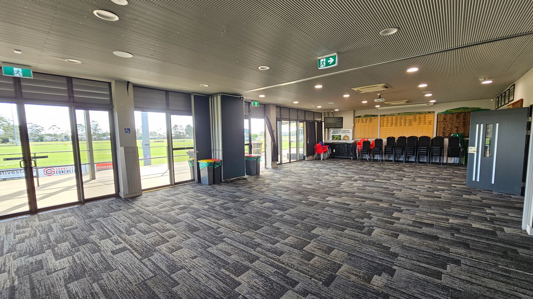 A spacious room with large glass doors on the left, offering a view of a grassy field outside. The floor is covered with patterned grey carpet tiles. On the right, there are stacked black chairs and a few red chairs near a table. The walls display wooden plaques. The ceiling is lined with recessed lights and features a grey, textured finish. An exit sign is visible above the doors. A spacious room with large glass doors on the left, offering a view of a grassy field outside. The floor is covered with patterned grey carpet tiles. On the right, there are stacked black chairs and a few red chairs near a table. The walls display wooden plaques. The ceiling is lined with recessed lights and features a grey, textured finish. An exit sign is visible above the doors.