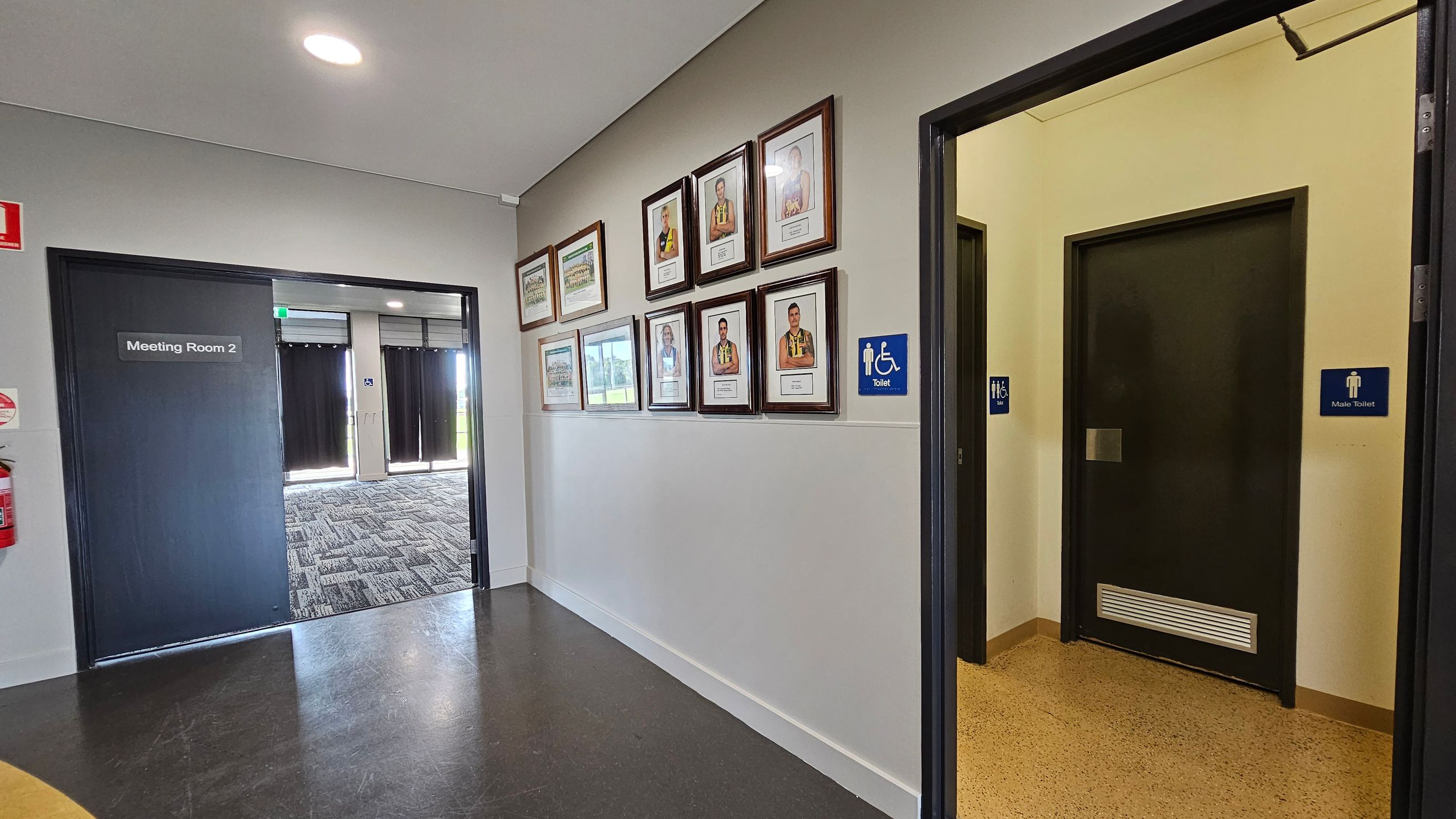 A hallway with a dark floor and light walls. On the left, there's a door labelled "Meeting Room 2" with a fire extinguisher nearby. The middle wall displays several framed photographs. On the right, there are two doors, one marked with a sign for an accessible toilet and the other for a male toilet. The ceiling is plain with a circular light fixture. A hallway with a dark floor and light walls. On the left, there's a door labelled "Meeting Room 2" with a fire extinguisher nearby. The middle wall displays several framed photographs. On the right, there are two doors, one marked with a sign for an accessible toilet and the other for a male toilet. The ceiling is plain with a circular light fixture.