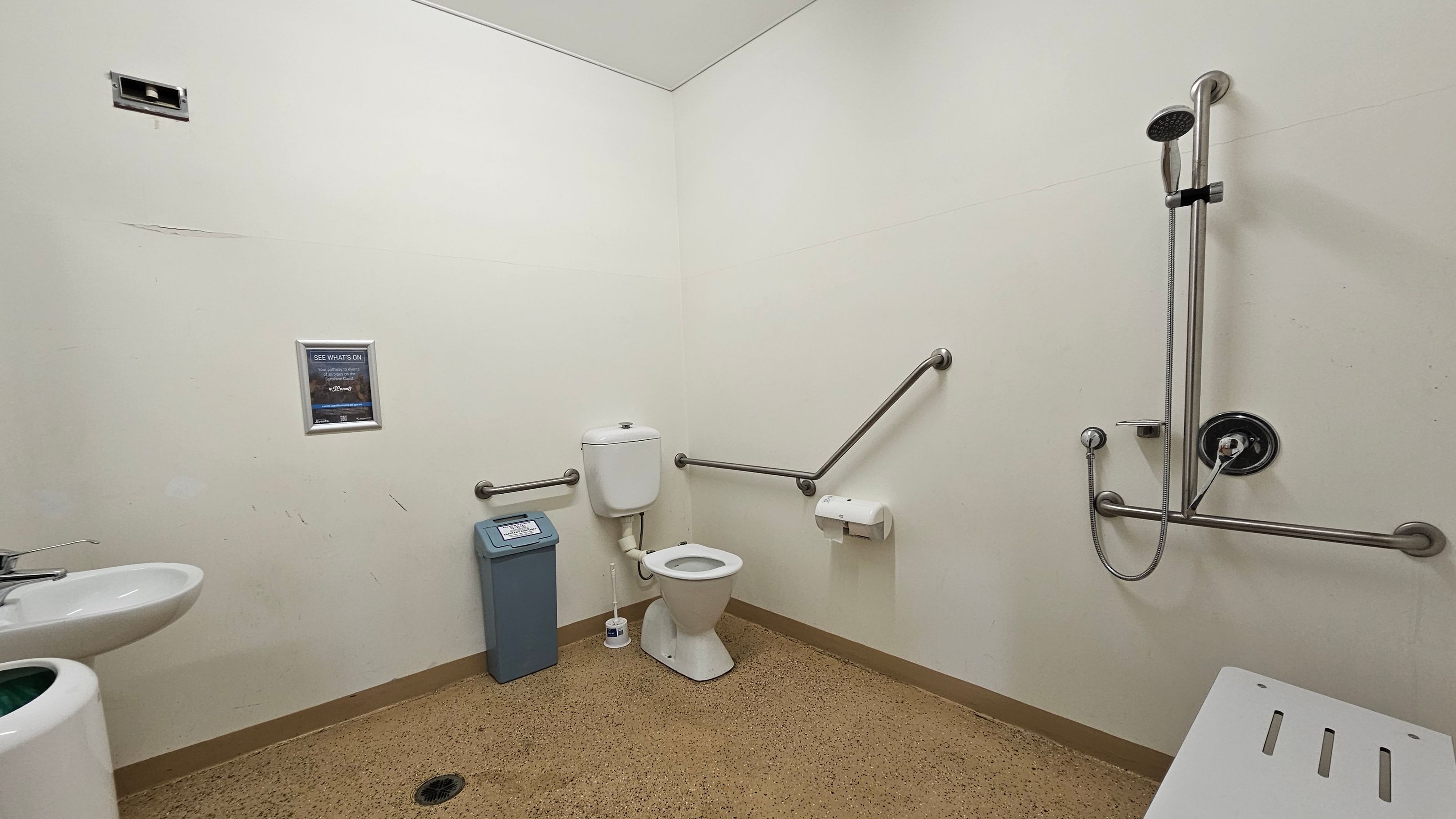 A bathroom with a white toilet in the centre, equipped with metal grab bars on both sides. To the left, there's a small sink with a tap and a waste bin nearby. On the right, a shower area with a handheld showerhead attached to a vertical rail and more grab bars. A white bench is partially visible in the foreground. The walls are light green, and the floor is speckled brown. A small poster is mounted on the wall above the bin. A bathroom with a white toilet in the centre, equipped with metal grab bars on both sides. To the left, there's a small sink with a tap and a waste bin nearby. On the right, a shower area with a handheld showerhead attached to a vertical rail and more grab bars. A white bench is partially visible in the foreground. The walls are light green, and the floor is speckled brown. A small poster is mounted on the wall above the bin.