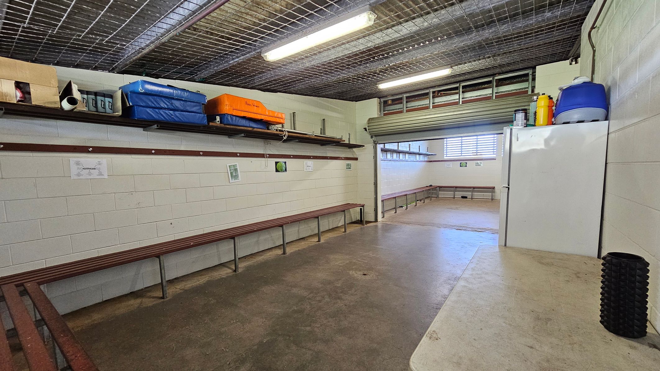 A room with concrete floors and a wire mesh ceiling. On the left, there are wooden benches along the walls and shelves above holding various items, including blue and orange mats. On the right, a white refrigerator with a blue cooler and yellow container on top. A black cylindrical object is on a nearby surface. The back wall has a partially open roller door and a window with horizontal bars, allowing light into the space. A room with concrete floors and a wire mesh ceiling. On the left, there are wooden benches along the walls and shelves above holding various items, including blue and orange mats. On the right, a white refrigerator with a blue cooler and yellow container on top. A black cylindrical object is on a nearby surface. The back wall has a partially open roller door and a window with horizontal bars, allowing light into the space.