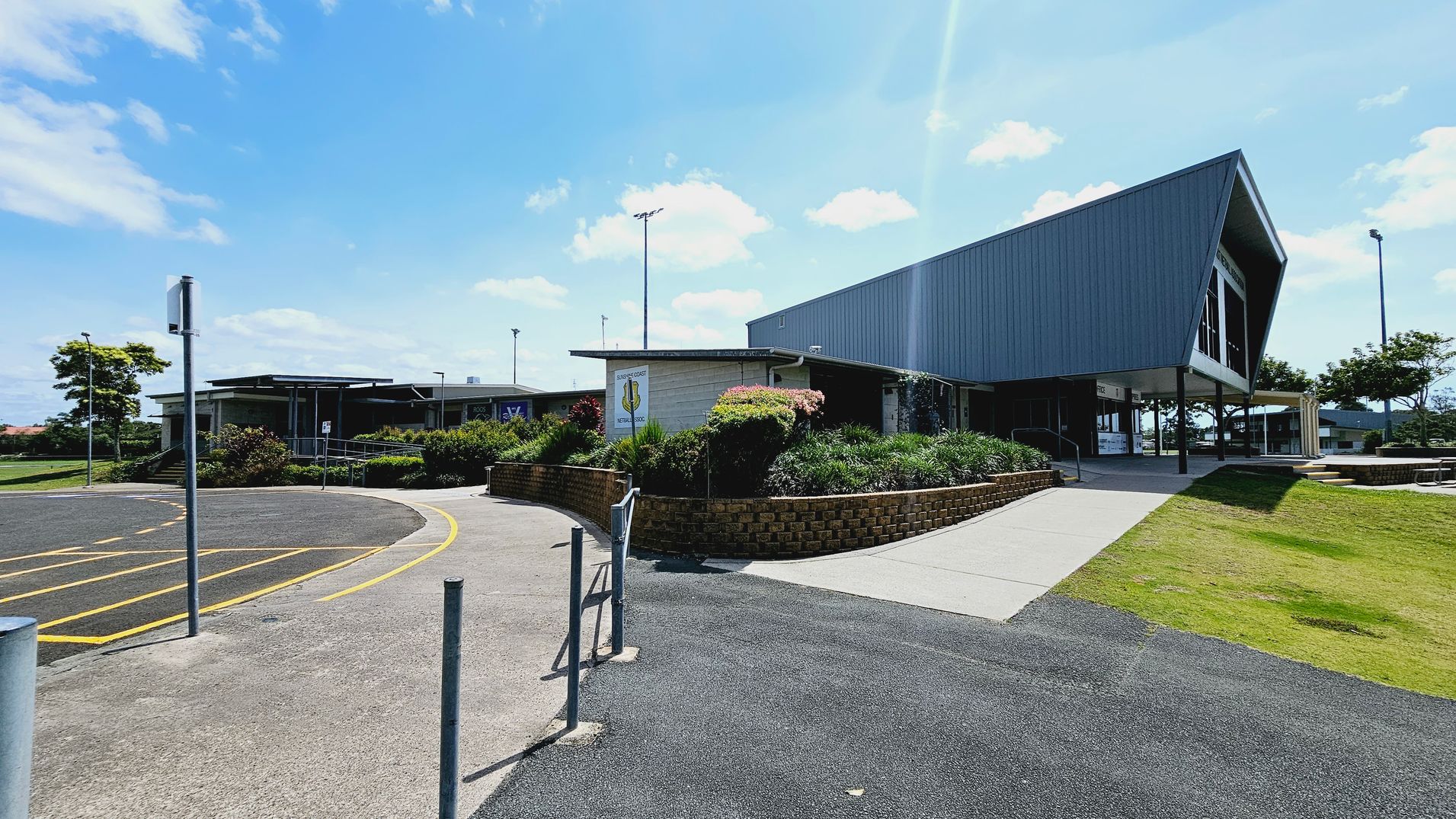 A modern building with a sign reading "Sunshine Coast Netball Association" is centred in the image. It has large windows and a flat roof. In front, there are several rows of metal picnic tables on tiered brick platforms. The foreground features a well-maintained grassy area with a concrete path leading to the building. On the left, there are power poles and a car park, while a large tree is visible on the right. A modern building with a sign reading "Sunshine Coast Netball Association" is centred in the image. It has large windows and a flat roof. In front, there are several rows of metal picnic tables on tiered brick platforms. The foreground features a well-maintained grassy area with a concrete path leading to the building. On the left, there are power poles and a car park, while a large tree is visible on the right.
