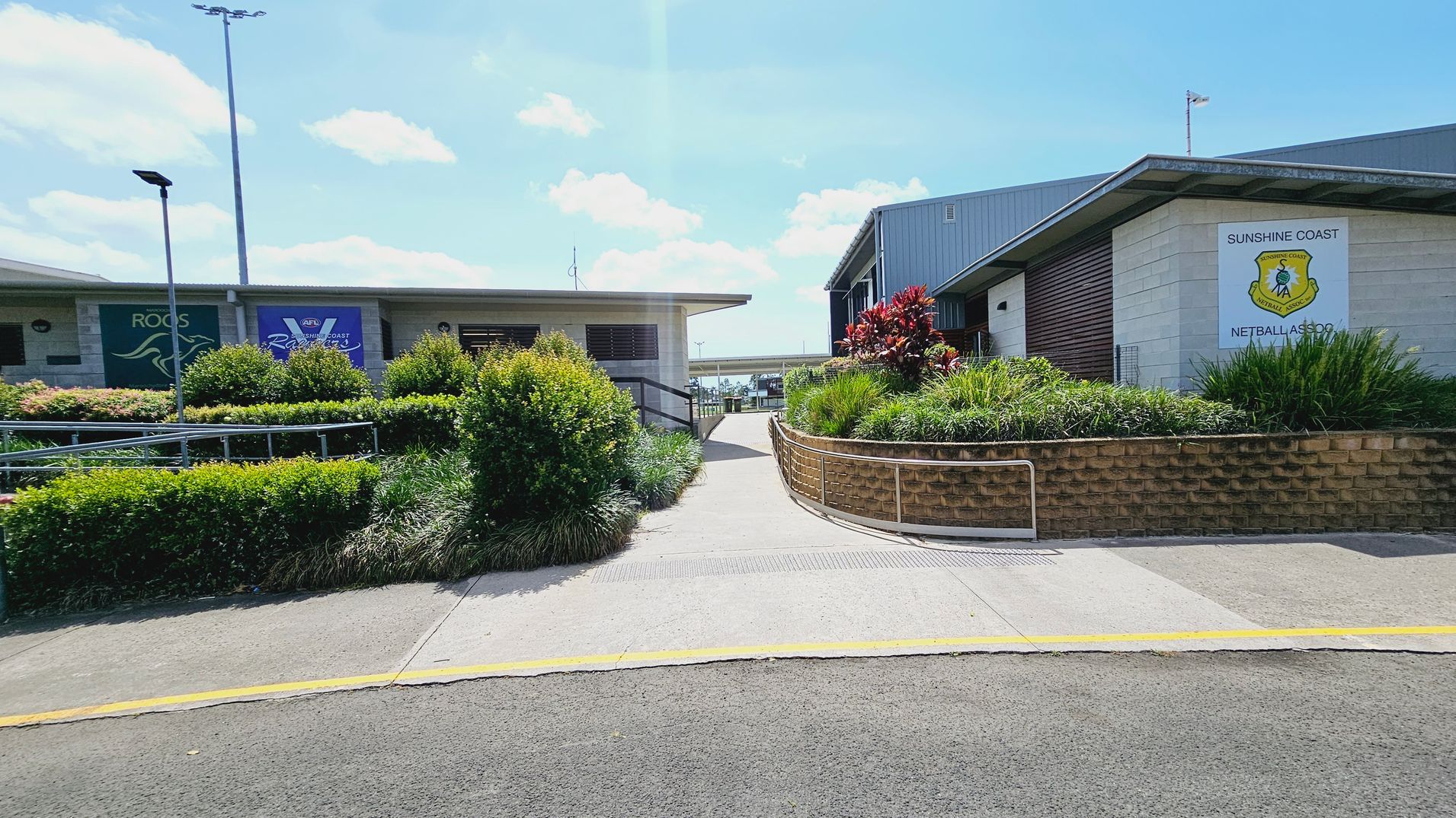 A pathway leads between two buildings. On the left, a building displays signs for "ROOS" and "AFL Sunshine Coast." On the right, another building has a sign for "Sunshine Coast Netball Association." Both buildings are surrounded by well-maintained greenery, including bushes and red-leaved plants. The path is bordered by a low brick wall and metal railing. The ground is paved, and there are tall light poles in the background. A pathway leads between two buildings. On the left, a building displays signs for "ROOS" and "AFL Sunshine Coast." On the right, another building has a sign for "Sunshine Coast Netball Association." Both buildings are surrounded by well-maintained greenery, including bushes and red-leaved plants. The path is bordered by a low brick wall and metal railing. The ground is paved, and there are tall light poles in the background.