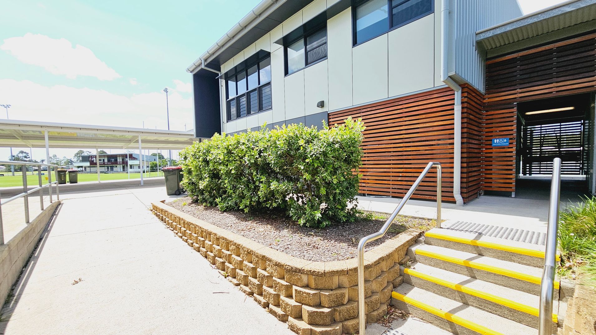 A modern building with a mix of metal and wooden panelling on the right. A set of concrete steps with yellow edges and metal railings leads up to an entrance. In the foreground, a raised garden bed with green shrubs is bordered by a brick wall. On the left, a covered walkway extends towards a sports field with a distant building. The ground is paved, and there are a few bins near the walkway. A modern building with a mix of metal and wooden panelling on the right. A set of concrete steps with yellow edges and metal railings leads up to an entrance. In the foreground, a raised garden bed with green shrubs is bordered by a brick wall. On the left, a covered walkway extends towards a sports field with a distant building. The ground is paved, and there are a few bins near the walkway.
