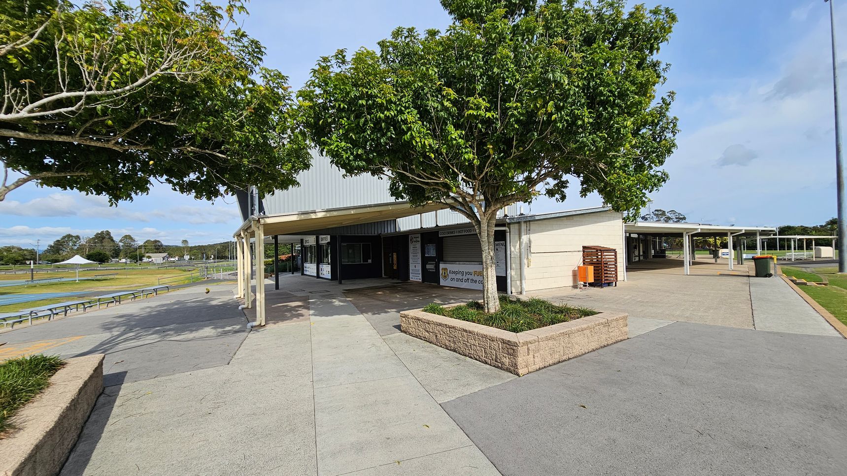 A paved outdoor area with a building in the centre, featuring a covered walkway. Two trees with lush green foliage are planted in raised brick planters. On the left, there are benches and a sports field with a white canopy in the distance. On the right, a pathway leads to more covered areas and a bin is visible. The ground is a mix of grey and green paving. A paved outdoor area with a building in the centre, featuring a covered walkway. Two trees with lush green foliage are planted in raised brick planters. On the left, there are benches and a sports field with a white canopy in the distance. On the right, a pathway leads to more covered areas and a bin is visible. The ground is a mix of grey and green paving.