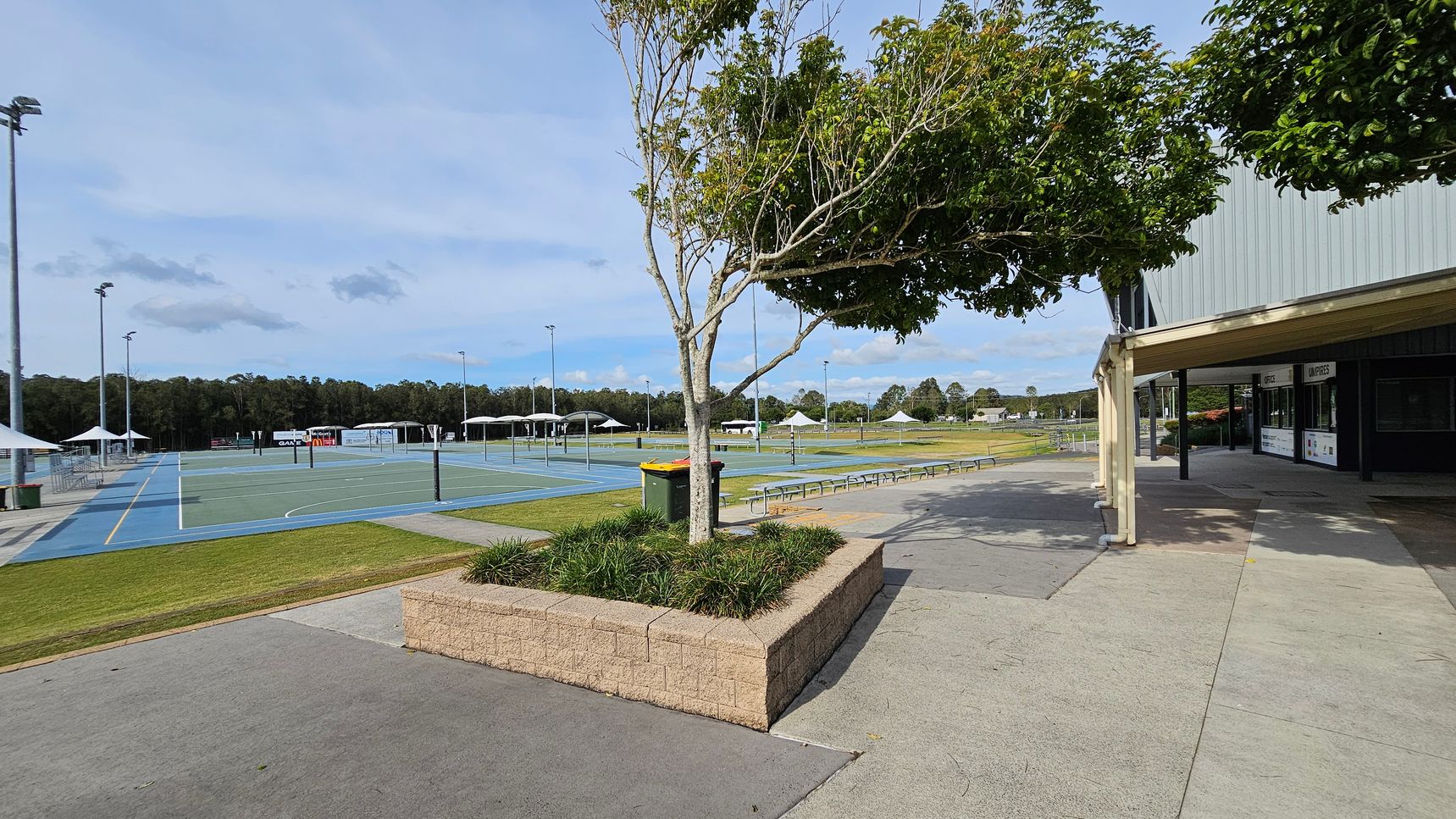 On the left, there are several outdoor sports courts with blue and green surfaces, surrounded by tall floodlights and white tents. In the middle, a tree is planted in a raised brick planter with grass. A green and yellow bin is nearby. On the right, a building with a green roof and a covered walkway is visible. The ground is a mix of concrete and grass, and there are benches along the path. In the background, there are trees and open fields. On the left, there are several outdoor sports courts with blue and green surfaces, surrounded by tall floodlights and white tents. In the middle, a tree is planted in a raised brick planter with grass. A green and yellow bin is nearby. On the right, a building with a green roof and a covered walkway is visible. The ground is a mix of concrete and grass, and there are benches along the path. In the background, there are trees and open fields.