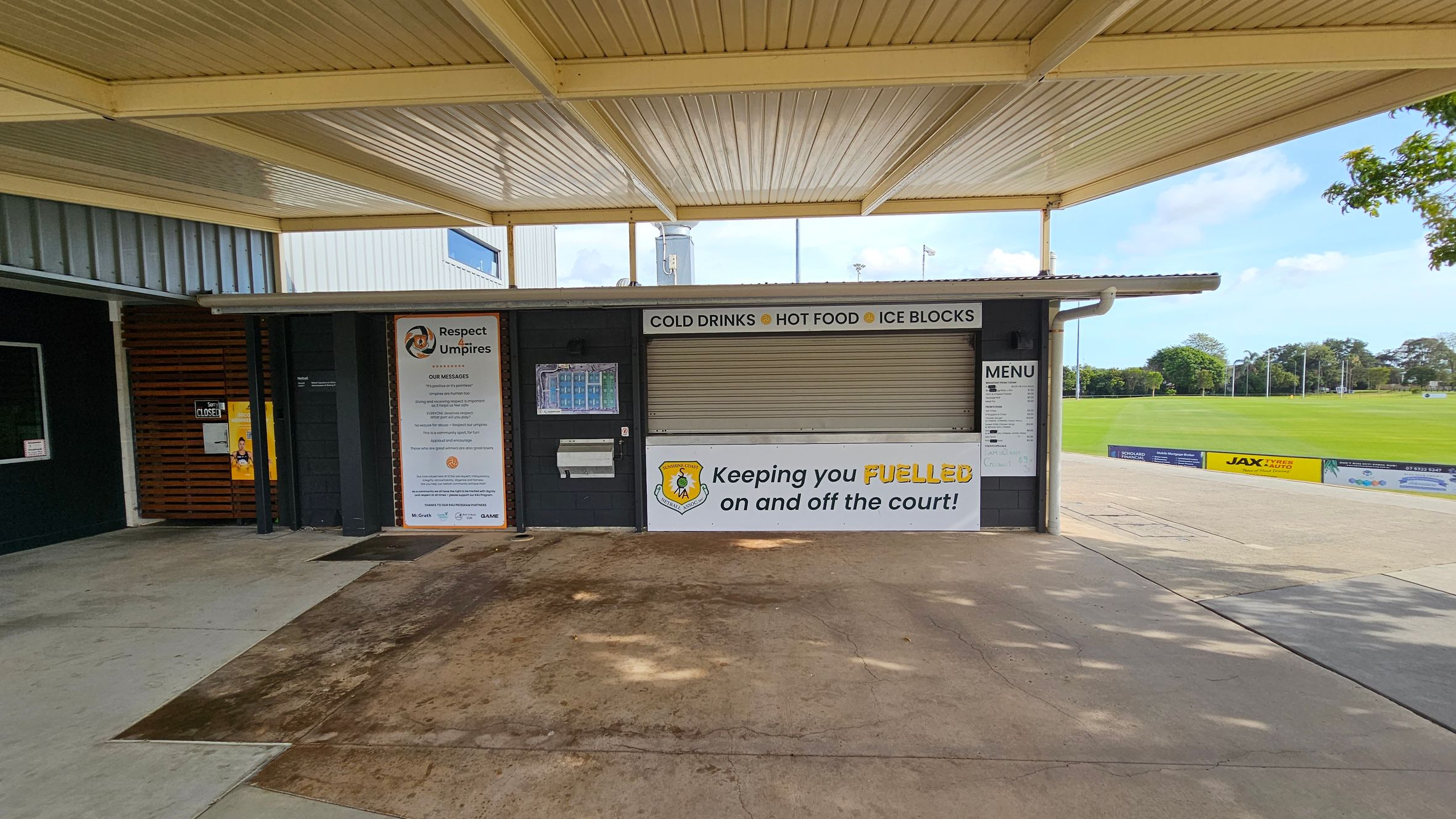 A closed kiosk with a corrugated metal roof and a concrete floor. The kiosk has a shuttered window with signs above it reading "COLD DRINKS," "HOT FOOD," and "ICE BLOCKS." A large sign on the left promotes respect for umpires. Another sign below the window says, "Keeping you FUELLED on and off the court!" A menu is displayed on the right. In the background, there is a grassy sports field with trees and a few advertisements along the fence. A closed kiosk with a corrugated metal roof and a concrete floor. The kiosk has a shuttered window with signs above it reading "COLD DRINKS," "HOT FOOD," and "ICE BLOCKS." A large sign on the left promotes respect for umpires. Another sign below the window says, "Keeping you FUELLED on and off the court!" A menu is displayed on the right. In the background, there is a grassy sports field with trees and a few advertisements along the fence.