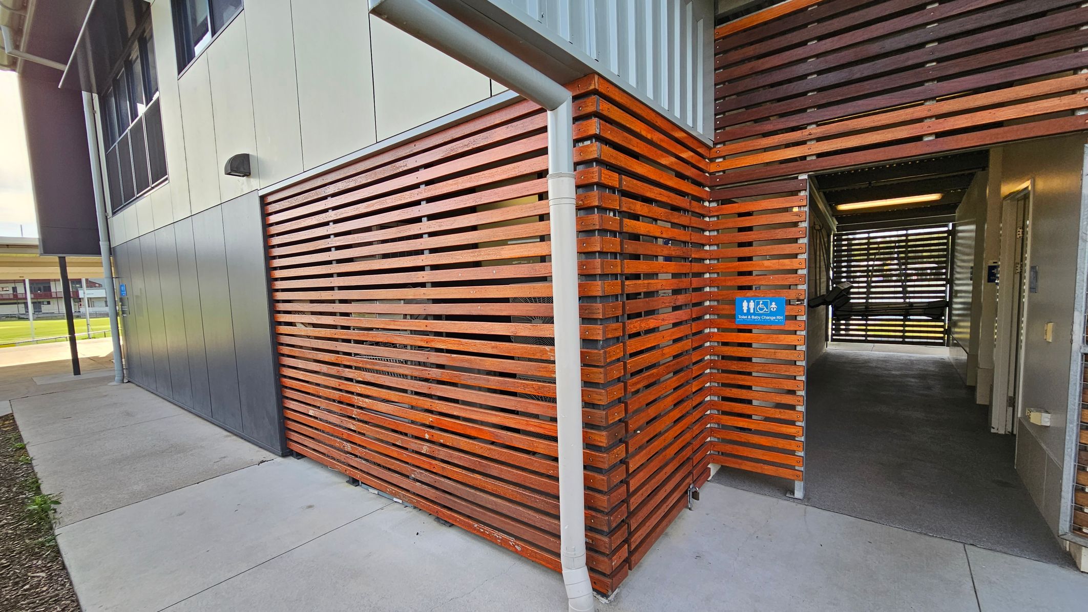 A modern building exterior with a combination of grey metal panels and horizontal wooden slats. The wooden slats form a corner and continue into a passageway. A white downpipe is attached to the corner. The entrance to the passageway has a blue sign indicating accessible toilets and baby changing facilities. The floor is concrete, and the interior passageway has a dark carpet. In the background, there is a glimpse of a grassy area and another building. A modern building exterior with a combination of grey metal panels and horizontal wooden slats. The wooden slats form a corner and continue into a passageway. A white downpipe is attached to the corner. The entrance to the passageway has a blue sign indicating accessible toilets and baby changing facilities. The floor is concrete, and the interior passageway has a dark carpet. In the background, there is a glimpse of a grassy area and another building.