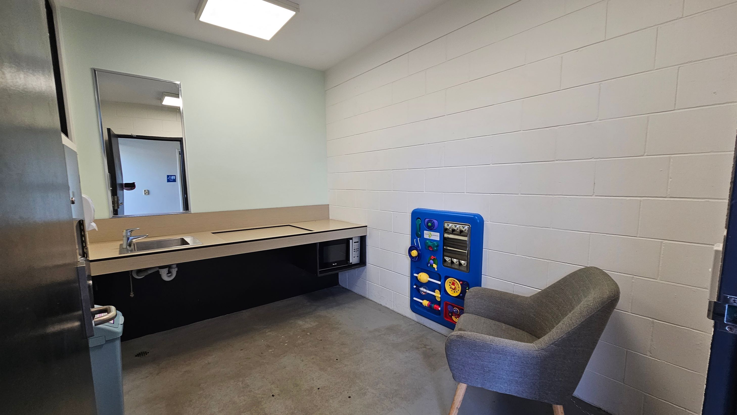 A small room with white brick walls and a grey concrete floor. On the left, there's a countertop with a sink and a microwave underneath. Above the counter is a mirror. On the right, a grey upholstered chair sits next to a blue activity panel attached to the wall. The ceiling has a square fluorescent light fixture. A small room with white brick walls and a grey concrete floor. On the left, there's a countertop with a sink and a microwave underneath. Above the counter is a mirror. On the right, a grey upholstered chair sits next to a blue activity panel attached to the wall. The ceiling has a square fluorescent light fixture.