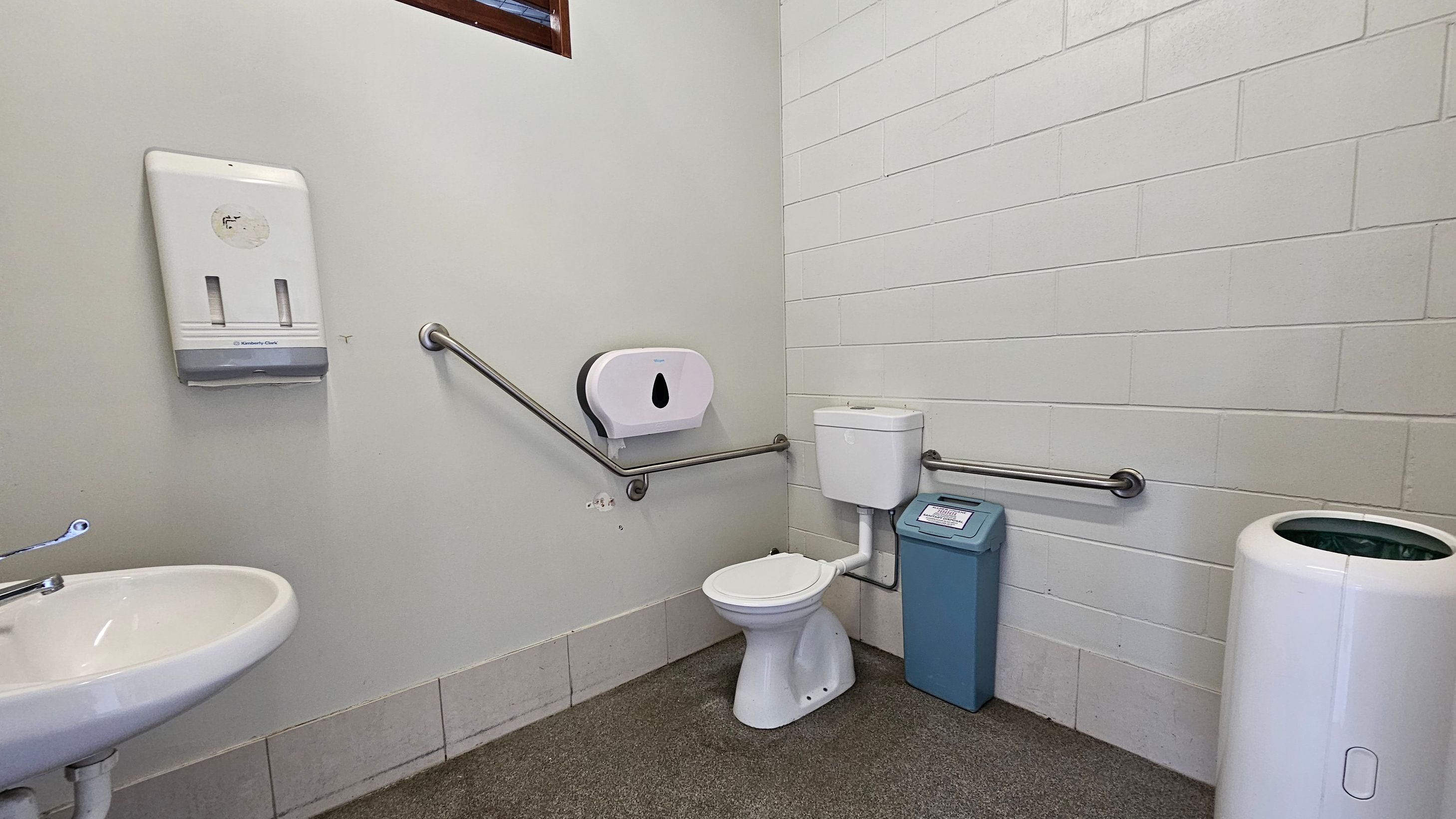 A public toilet with a white toilet bowl and cistern in the centre, equipped with metal grab bars on both sides. To the left, a white sink with a metal tap is visible, along with a wall-mounted paper towel dispenser. On the right, a blue sanitary bin and a white waste bin are present. The walls are tiled in white, and the floor is covered with a mix of grey and brown tiles. A public toilet with a white toilet bowl and cistern in the centre, equipped with metal grab bars on both sides. To the left, a white sink with a metal tap is visible, along with a wall-mounted paper towel dispenser. On the right, a blue sanitary bin and a white waste bin are present. The walls are tiled in white, and the floor is covered with a mix of grey and brown tiles.