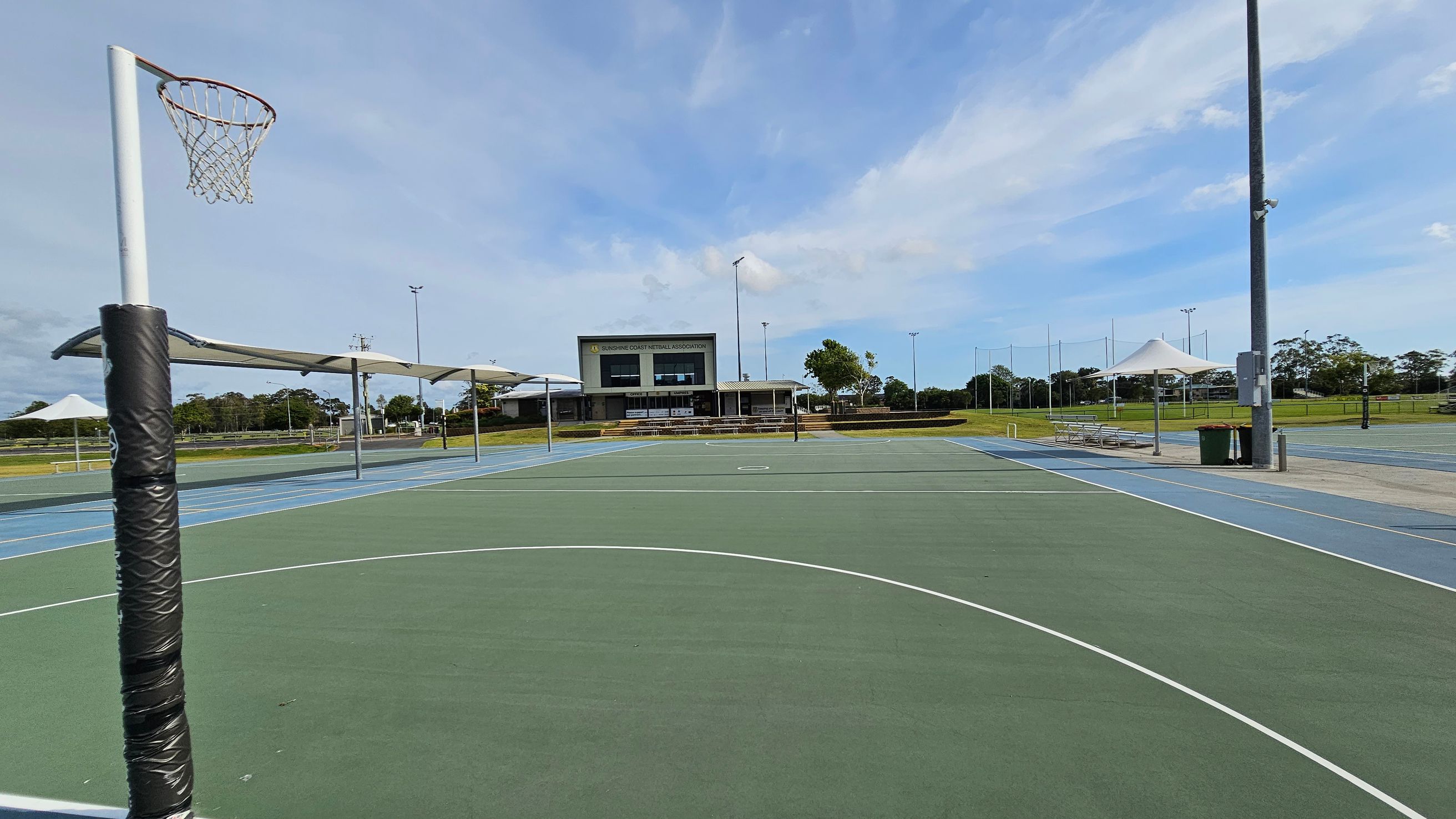 Outdoor netball courts with a green playing surface and white lines. A netball hoop with a padded post is on the left. In the background, there is a building with a sign, "Sunshine Coast Netball Association." Several covered seating areas and a rubbish bin are visible on the right. The area is surrounded by grass and trees, with tall light poles around the courts. Outdoor netball courts with a green playing surface and white lines. A netball hoop with a padded post is on the left. In the background, there is a building with a sign, "Sunshine Coast Netball Association." Several covered seating areas and a rubbish bin are visible on the right. The area is surrounded by grass and trees, with tall light poles around the courts.