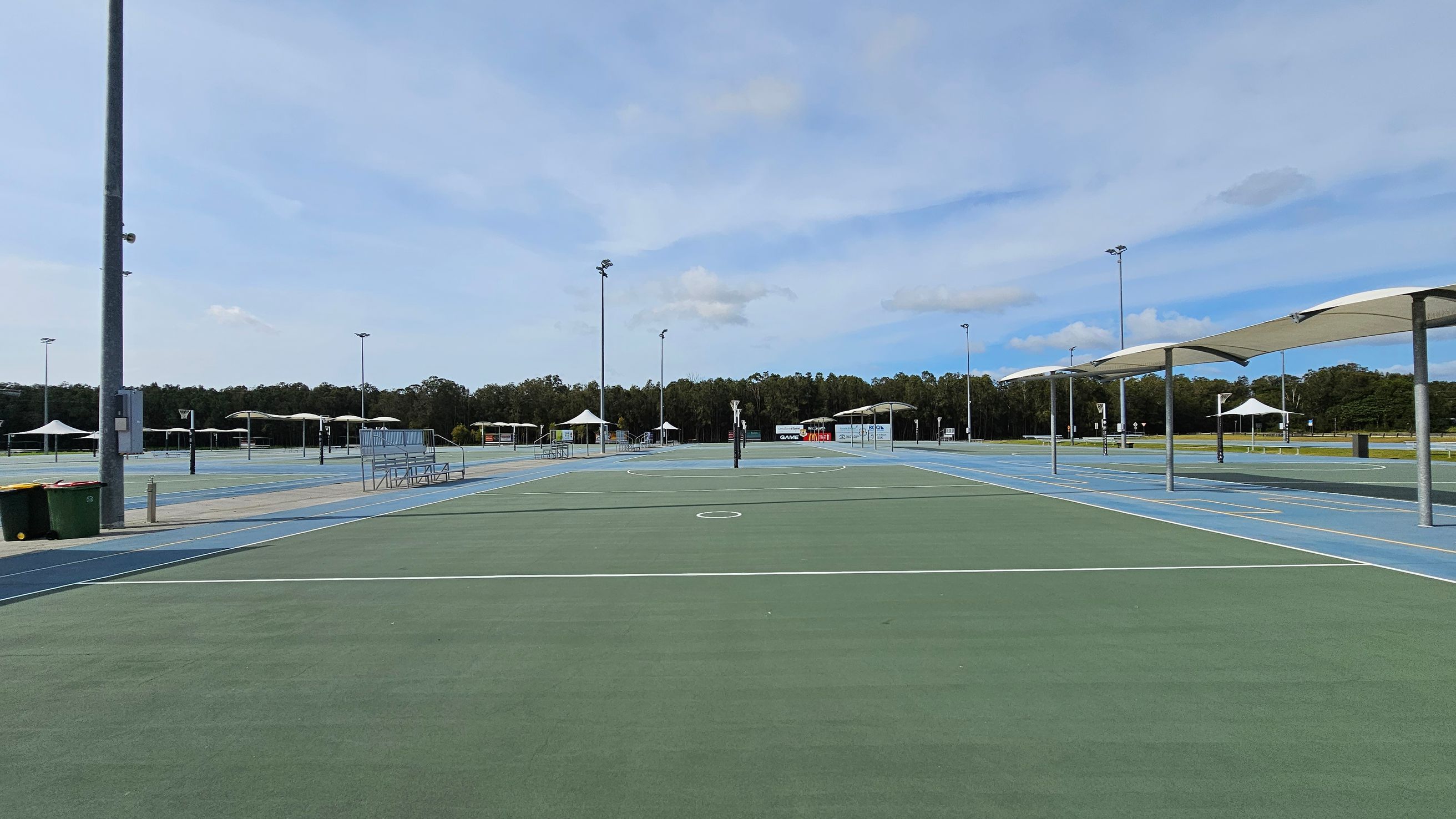 A large outdoor sports complex with multiple netball courts. The courts have green and blue surfaces with white lines. On the left, there are bins and a tall light pole. In the centre, a netball post is visible. To the right, there are covered seating areas with white canopies. In the background, there are more courts, additional canopies, and a line of trees. A large outdoor sports complex with multiple netball courts. The courts have green and blue surfaces with white lines. On the left, there are bins and a tall light pole. In the centre, a netball post is visible. To the right, there are covered seating areas with white canopies. In the background, there are more courts, additional canopies, and a line of trees.