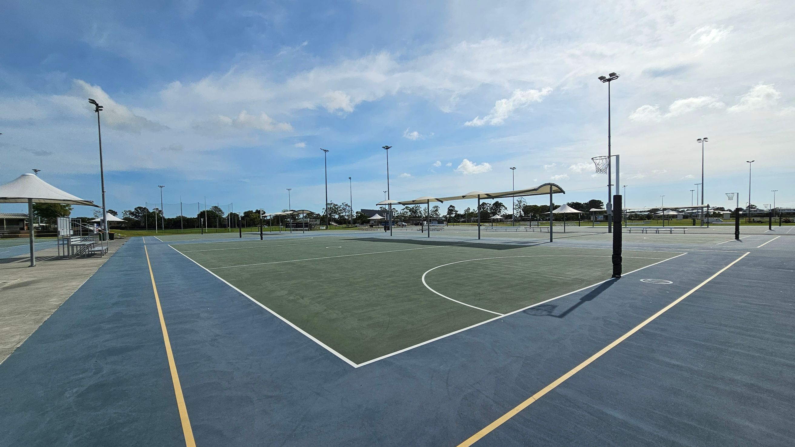 Outdoor netball courts with blue and green surfaces, marked with yellow and white lines. On the left, a covered seating area with a white canopy and metal benches. Several tall floodlights are positioned around the courts. In the background, there are more courts and some trees. The area is open and spacious, with clear visibility of the surrounding landscape. Outdoor netball courts with blue and green surfaces, marked with yellow and white lines. On the left, a covered seating area with a white canopy and metal benches. Several tall floodlights are positioned around the courts. In the background, there are more courts and some trees. The area is open and spacious, with clear visibility of the surrounding landscape.