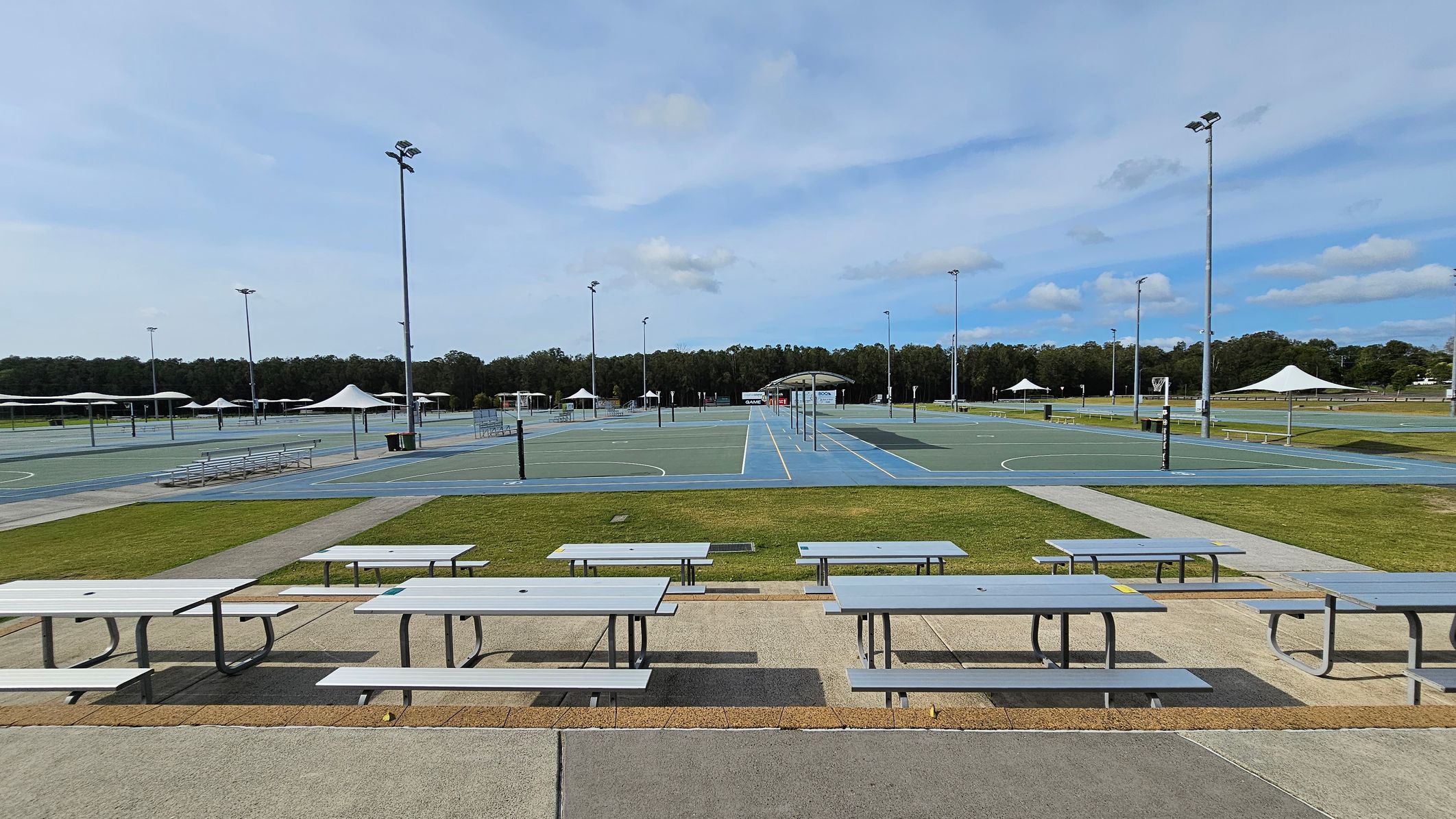 Rows of light grey picnic tables are in the foreground on a paved area. Beyond them, there are multiple outdoor sports courts with blue and green surfaces, surrounded by tall floodlights. White shade structures are scattered around the courts. In the background, there is a line of trees bordering the area. The ground is a mix of grass and concrete paths. Rows of light grey picnic tables are in the foreground on a paved area. Beyond them, there are multiple outdoor sports courts with blue and green surfaces, surrounded by tall floodlights. White shade structures are scattered around the courts. In the background, there is a line of trees bordering the area. The ground is a mix of grass and concrete paths.